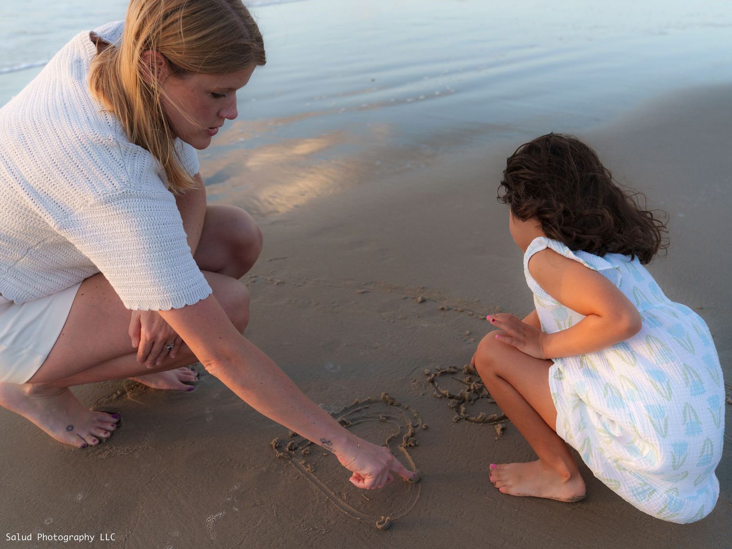 Person in white dress crouches to write in sand at water's edge.