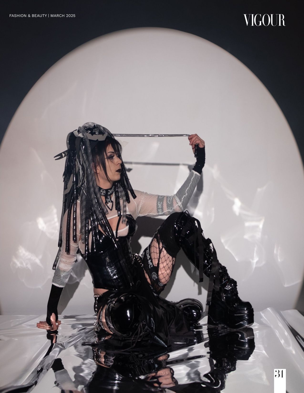 Dark gothic fashion model in boots and black lace poses dramatically on reflective white surface in editorial photoshoot.