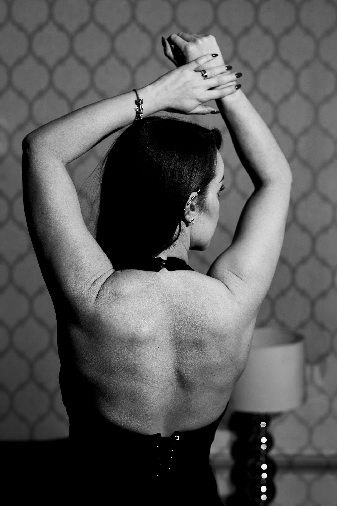 Black and white photograph showing the muscular back of a person with raised arms against a patterned wall.