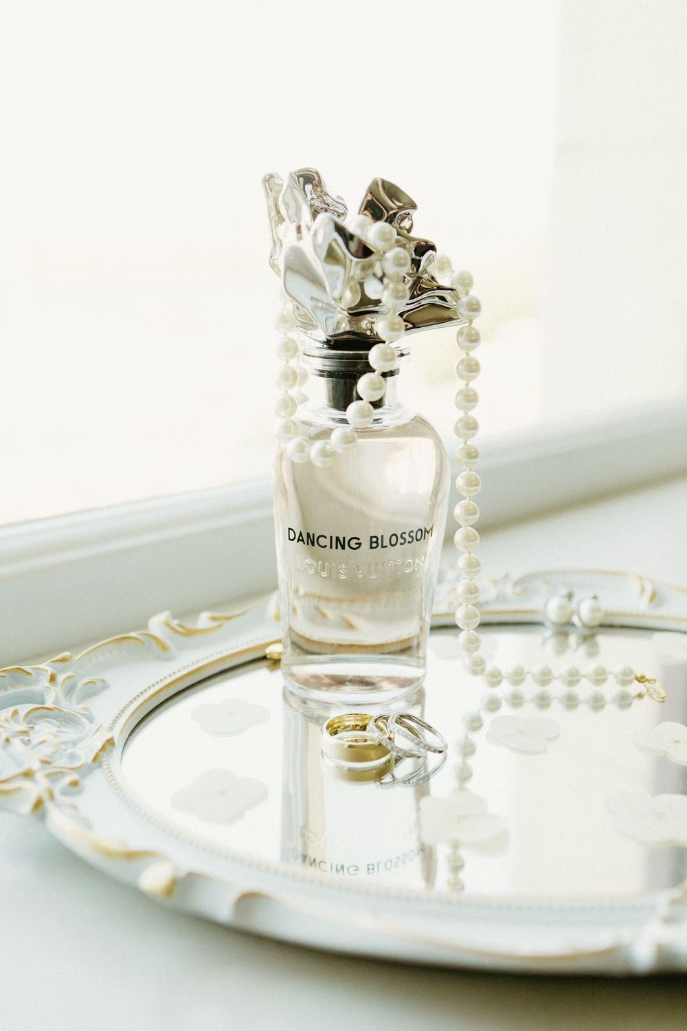 Elegant perfume bottle and pearl necklace arranged on silver tray in soft light.