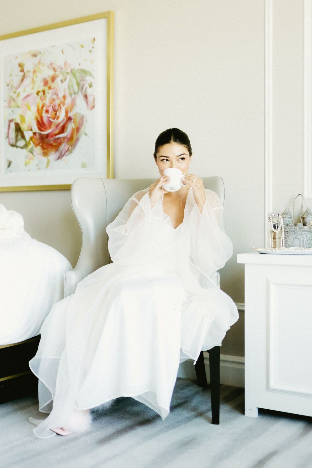 A figure in a flowing white dress sits elegantly in a bright room with floral artwork on the walls.