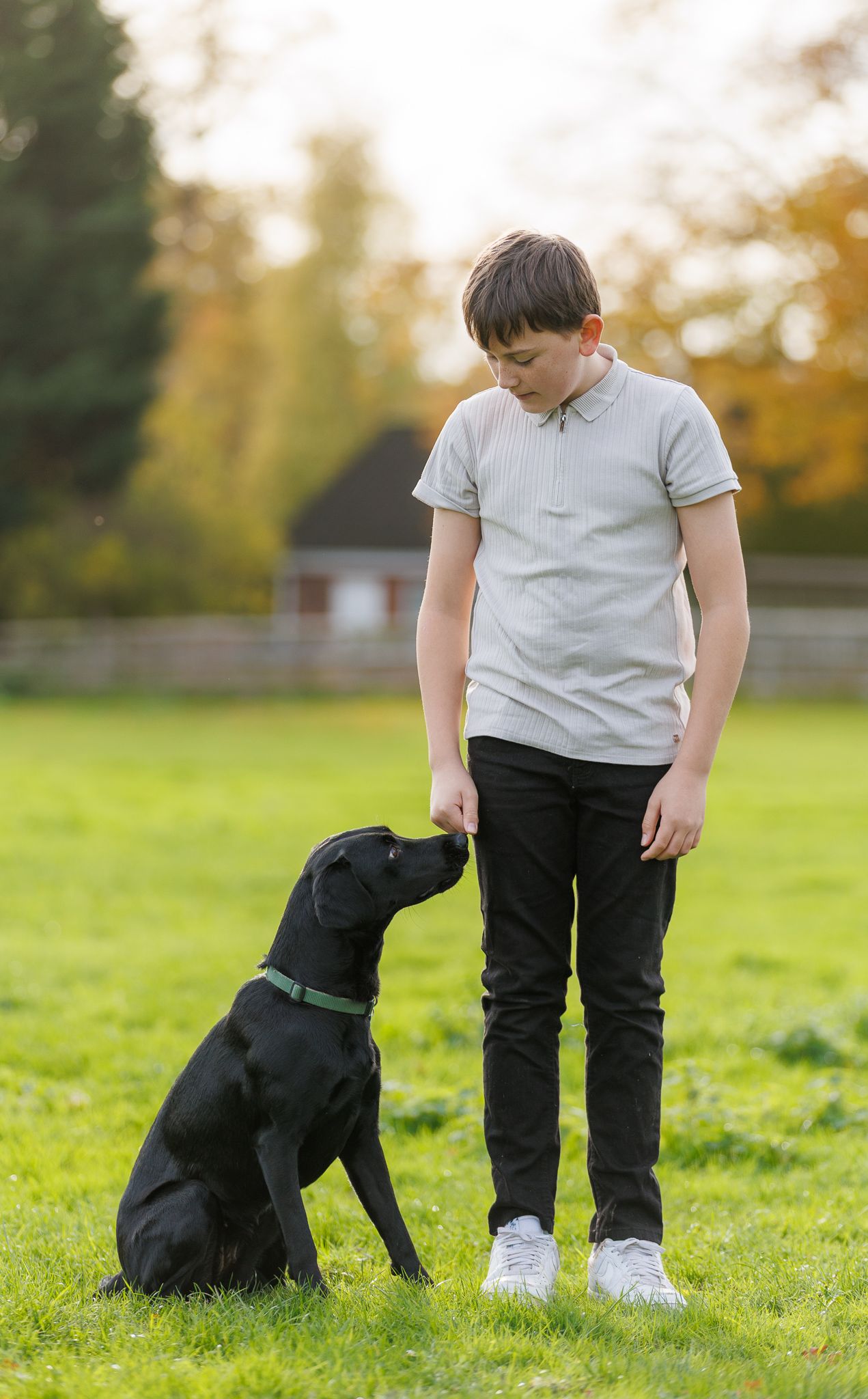 A black dog sits obediently next to a person in a white shirt and dark pants on a grassy field during autumn.
