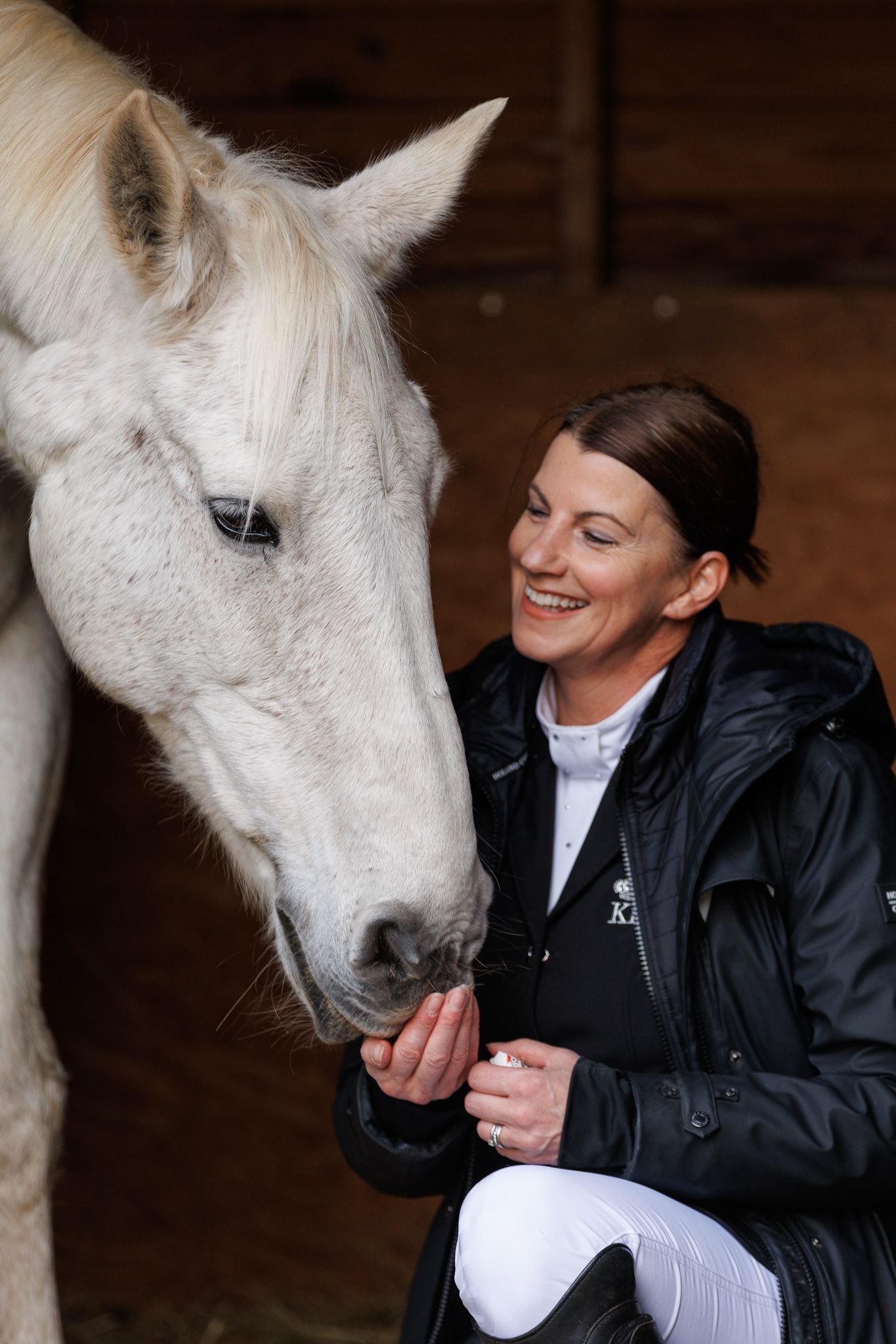 A beautiful white horse and a person in a black jacket share a tender moment in a stable.