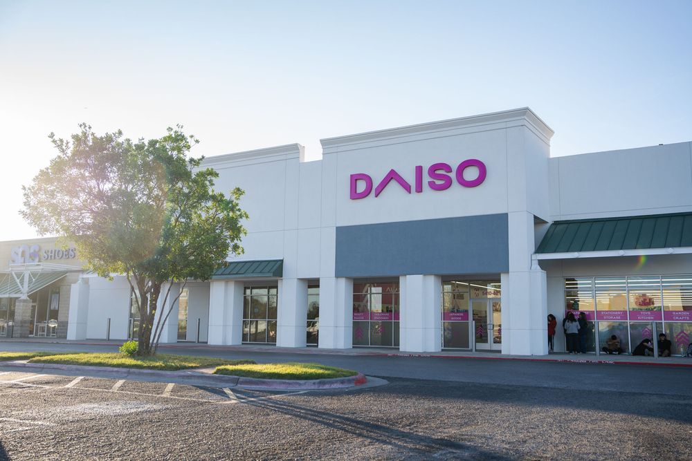 A strip mall exterior with white storefronts and a Daiso retail store sign visible against a blue sky.