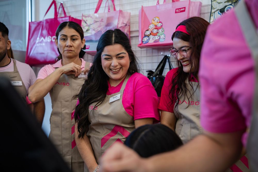 Group of retail employees wearing pink ribbons and tan uniforms smile together in store aisle.