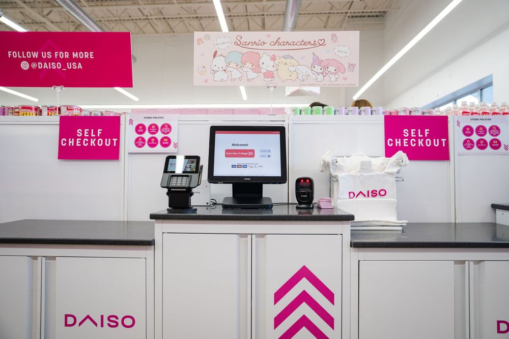 Interior view of a Daiso retail store with bright pink signage and white walls showing checkout counters and displays.