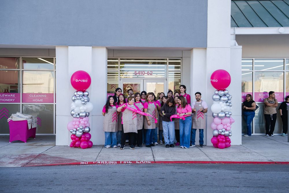 A group of people stand outside a storefront entrance decorated with pink balloons and signage for a grand opening event.