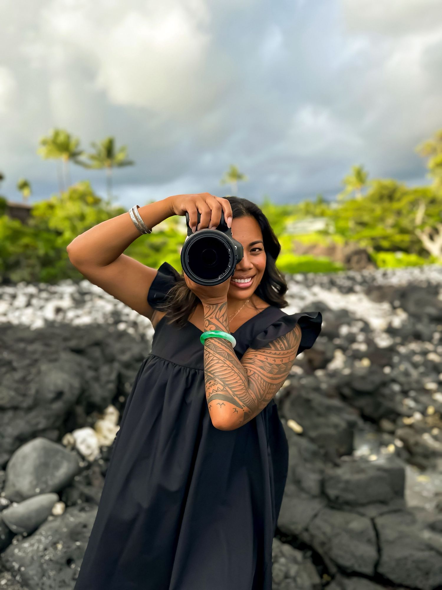 Photographer with intricate arm tattoo takes a photo on rocky volcanic beach with palm trees and cloudy sky in Hawaii.