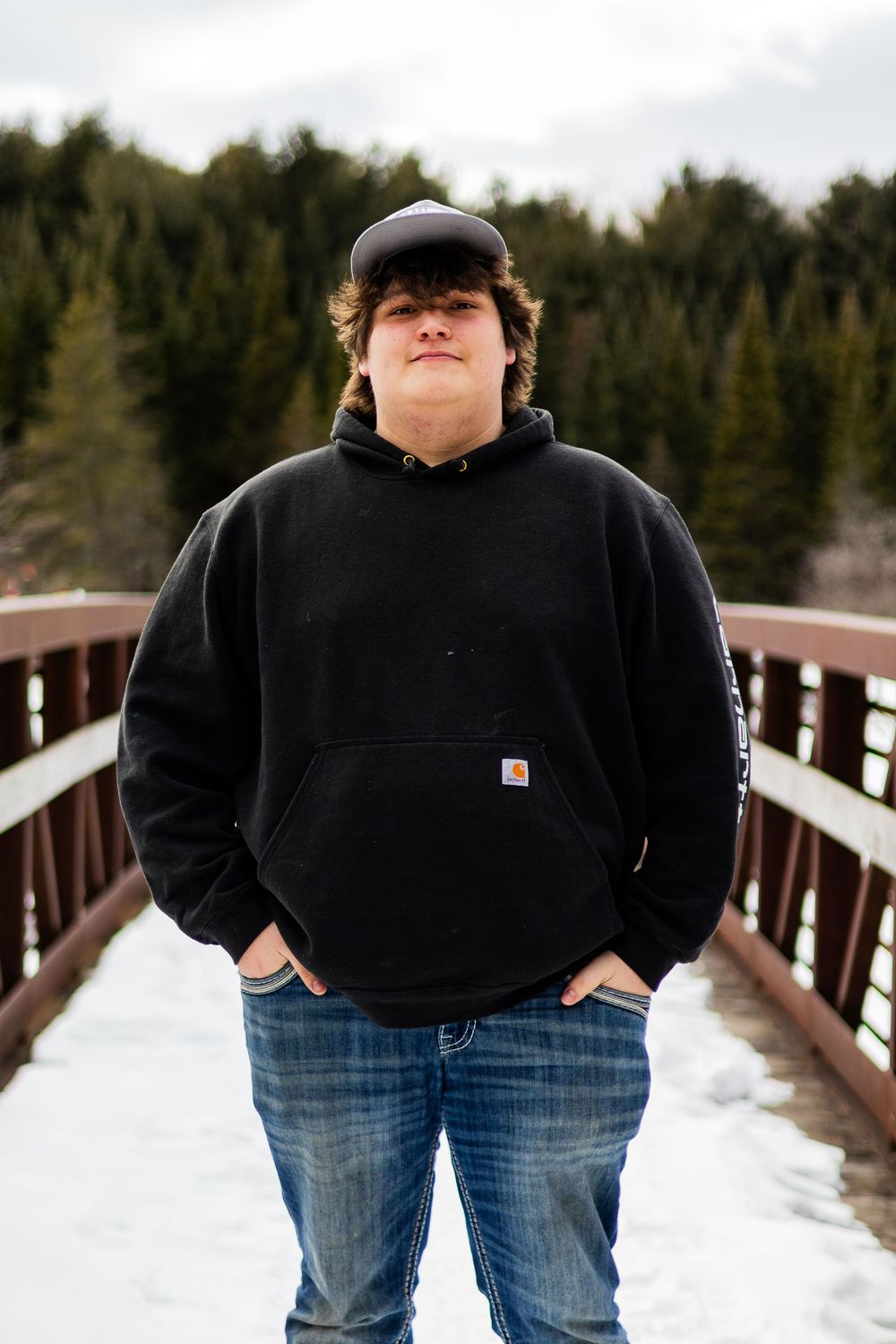 Person in black hoodie standing on wooden walkway surrounded by snow and evergreens.