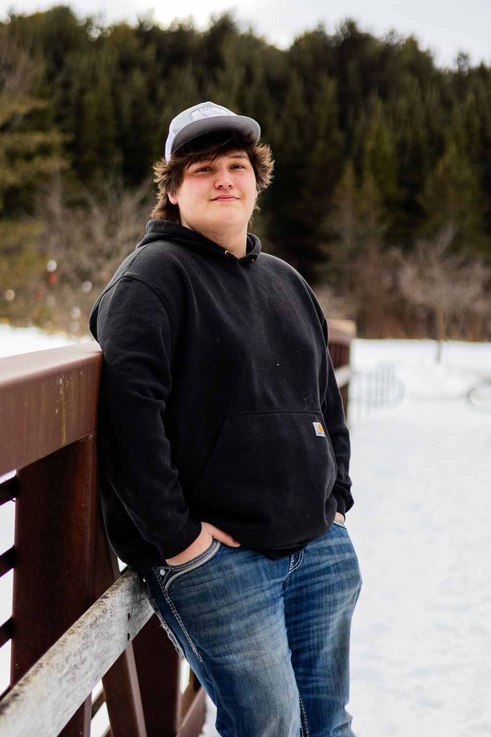 Person wearing black hoodie and jeans poses against snowy winter backdrop on wooden bridge.