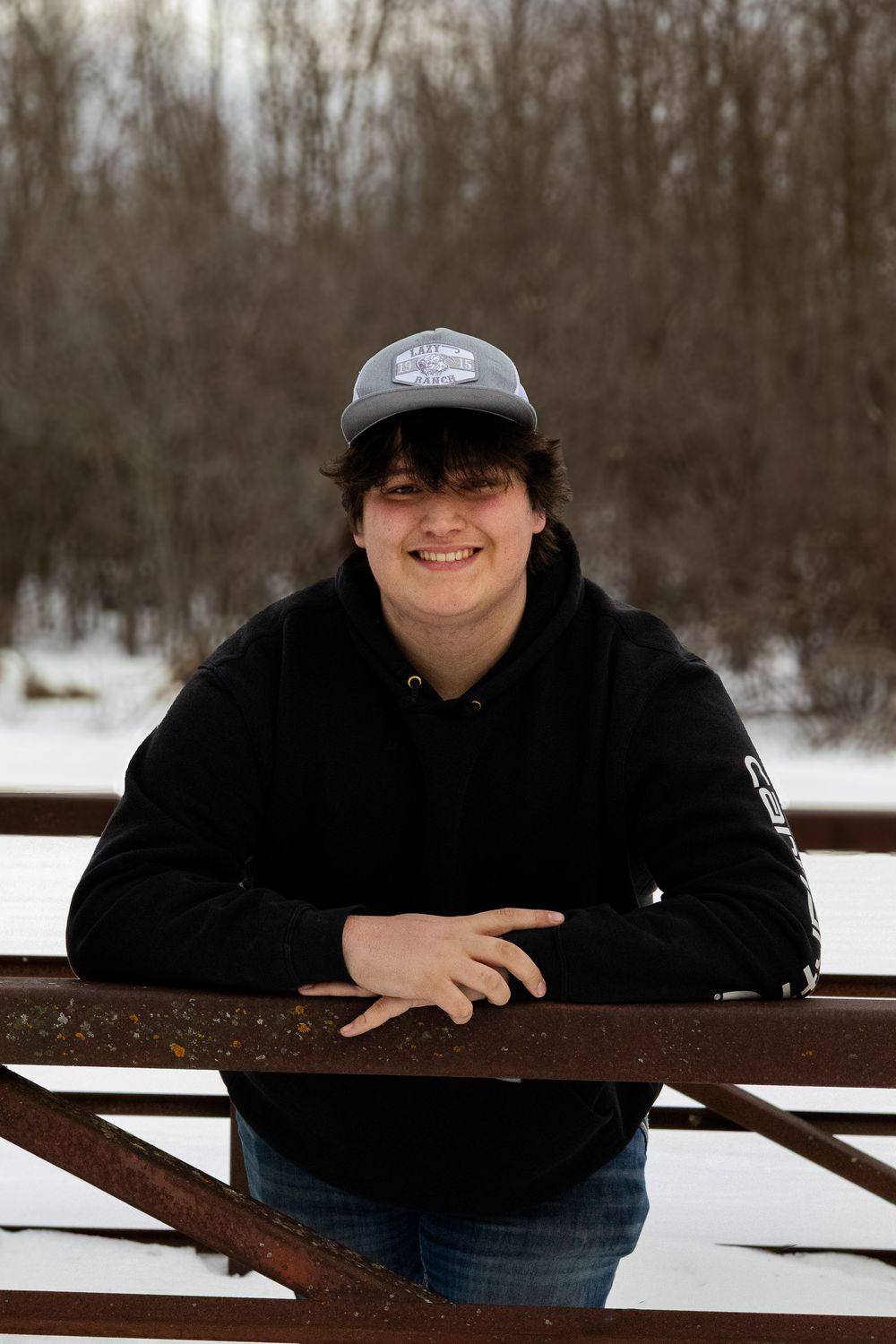Person in baseball cap and black hoodie leans on wooden railing in winter setting.