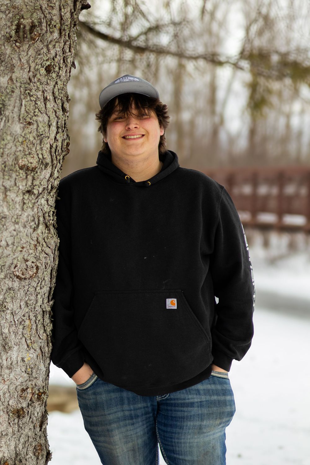 Figure in Carhartt hoodie and jeans stands between snow-covered trees in winter landscape.
