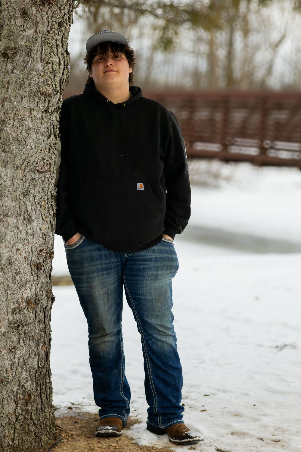 Person in casual winter attire poses near tree trunk in snowy outdoor setting.