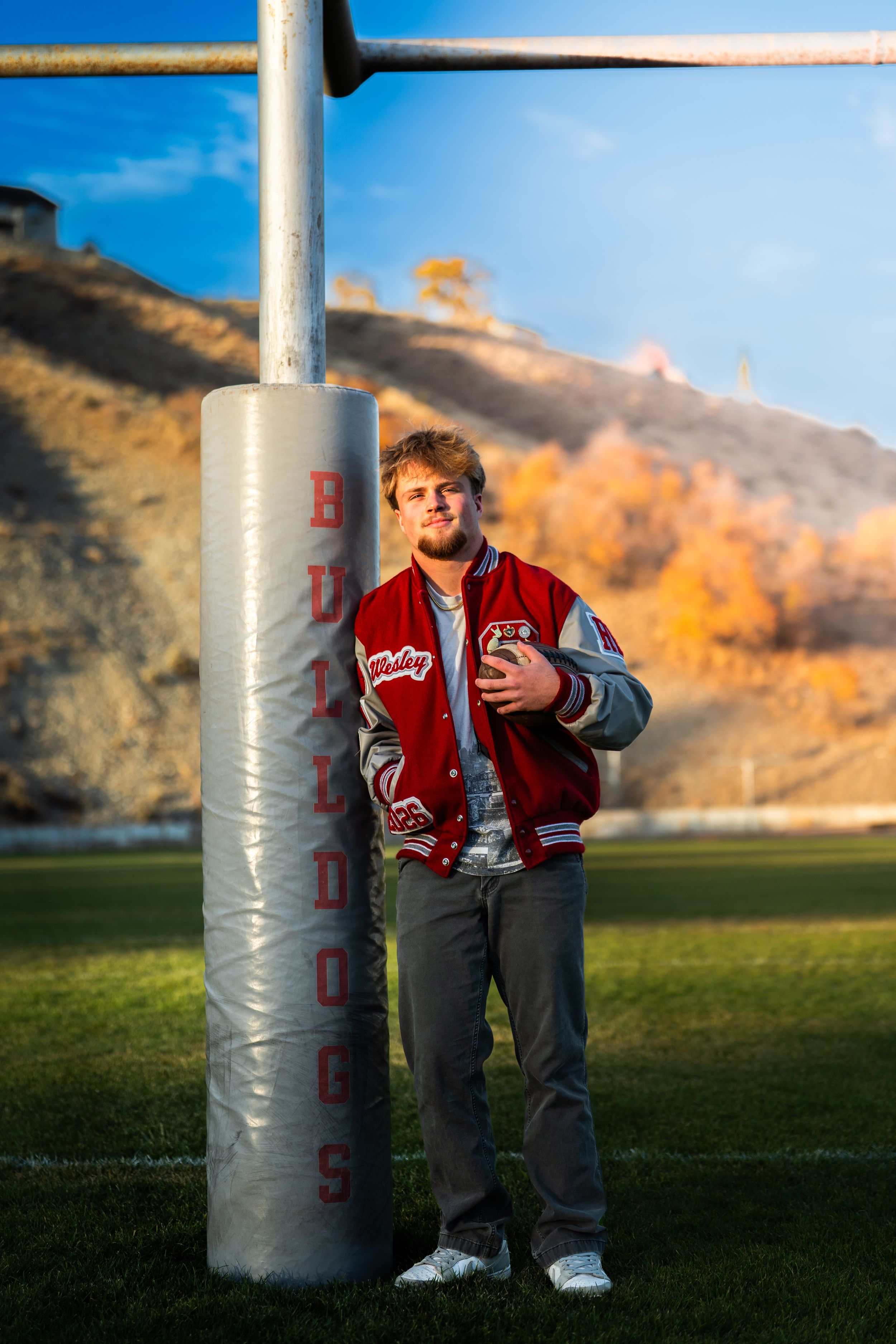 Athletic figure in varsity jacket leaning against football goal post during autumn.