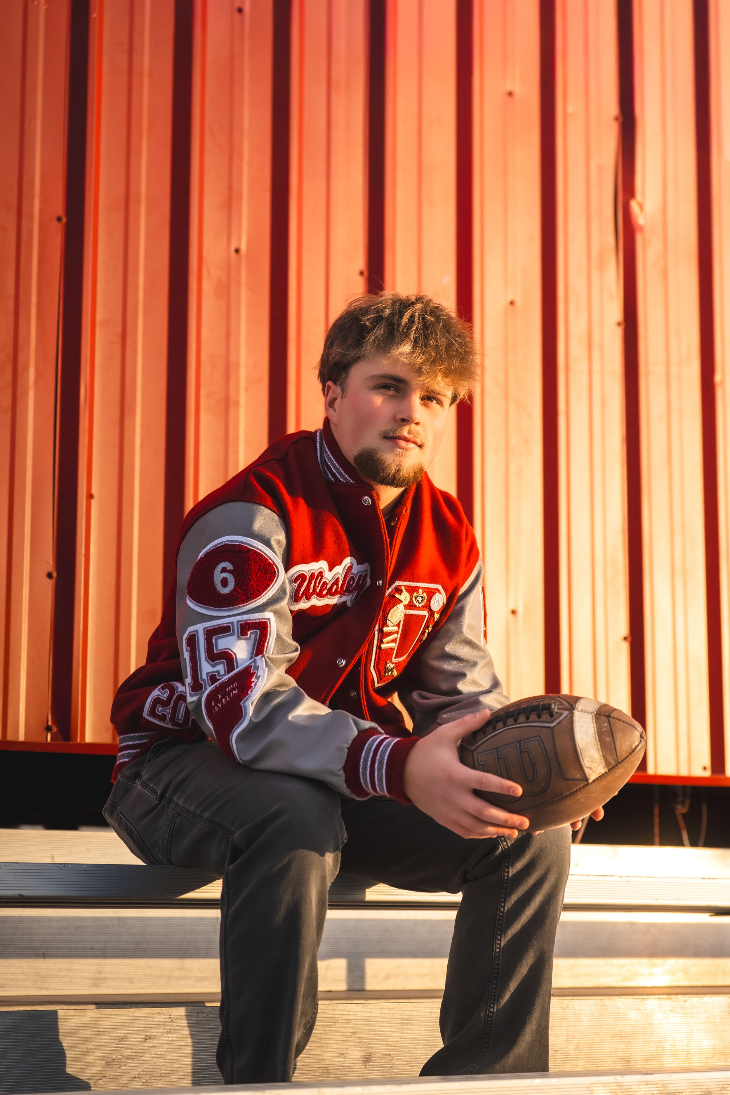 Student athlete in red jersey sitting on bench with football at sunset.