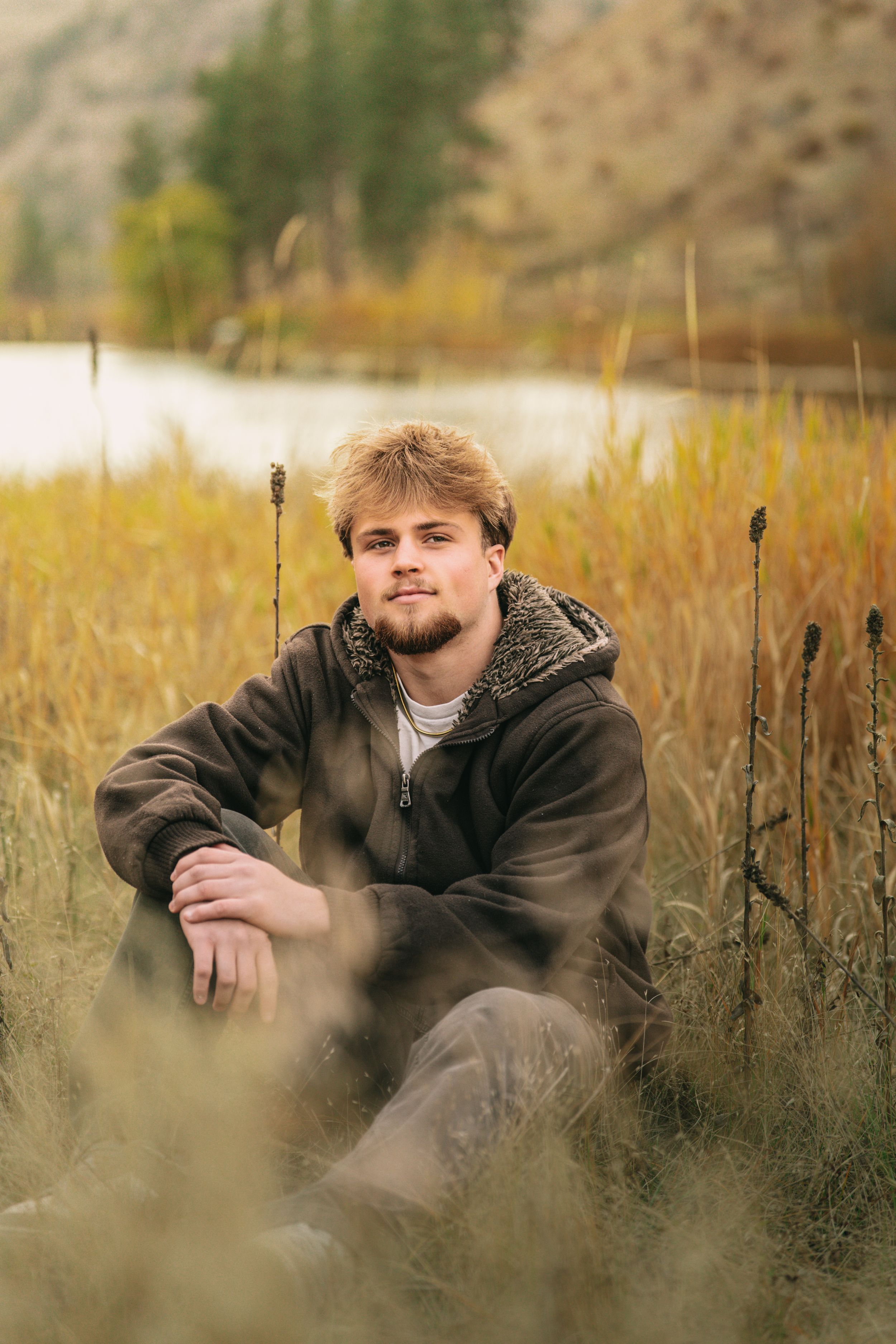 Person in brown jacket sitting in tall autumn grass during golden hour photography session.