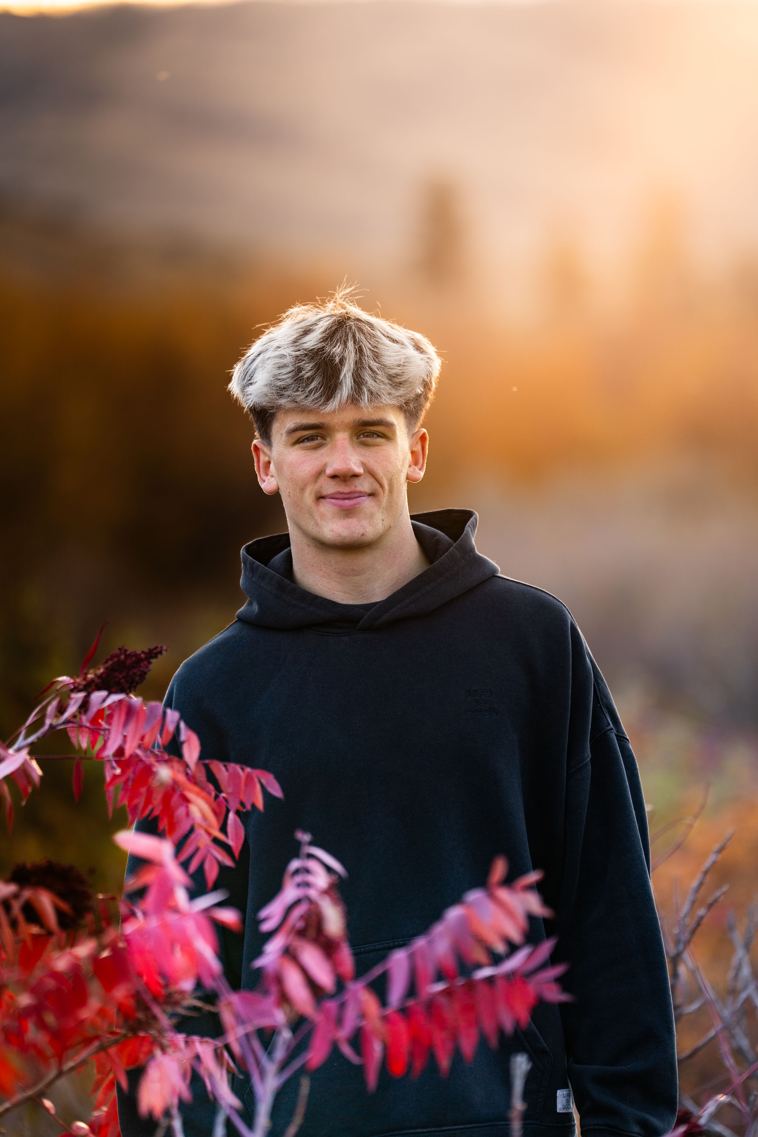 Person wearing black hoodie smiling in front of vibrant red autumn foliage at sunset.