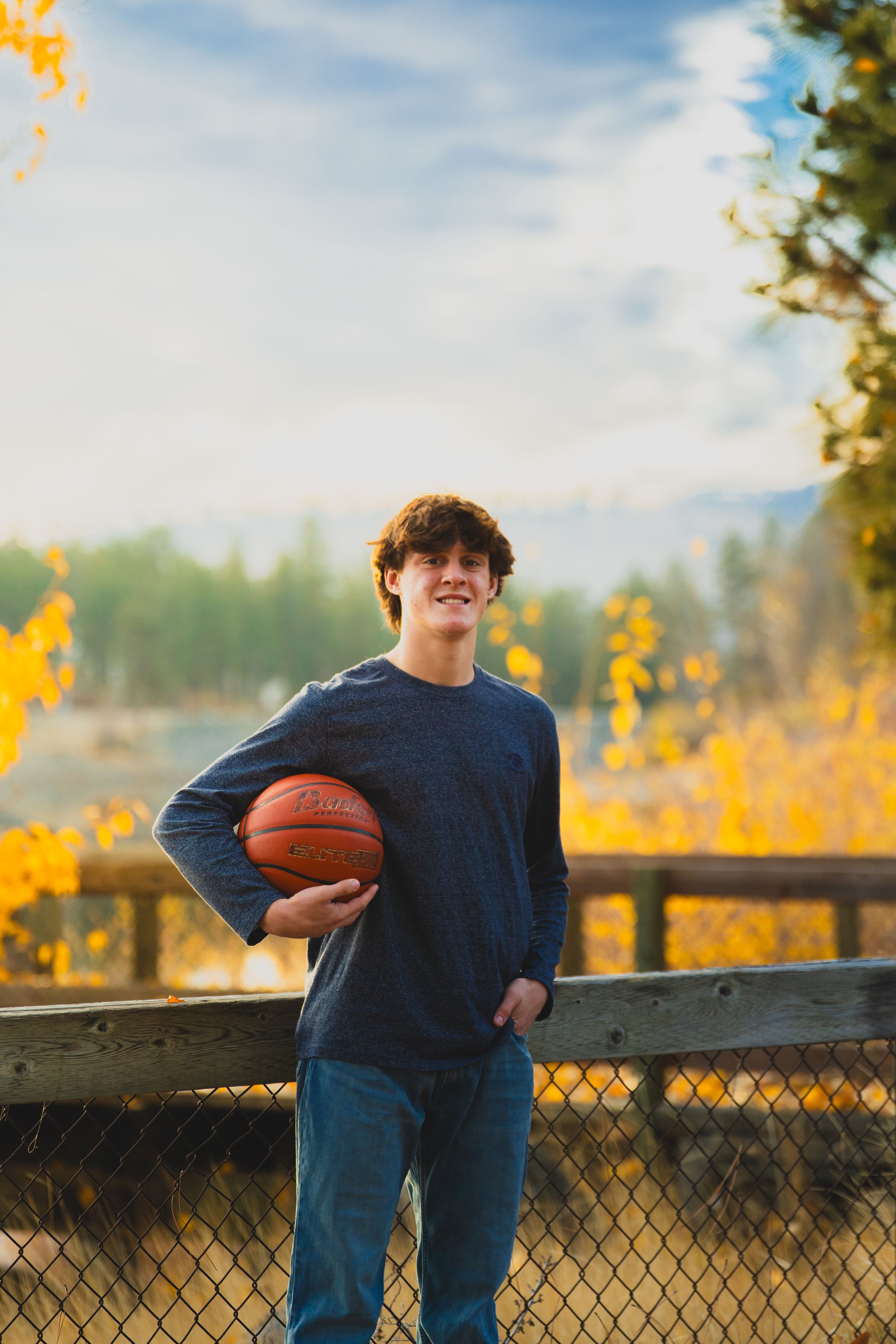 Athletic figure in blue holding a basketball against autumn foliage and mountain backdrop.