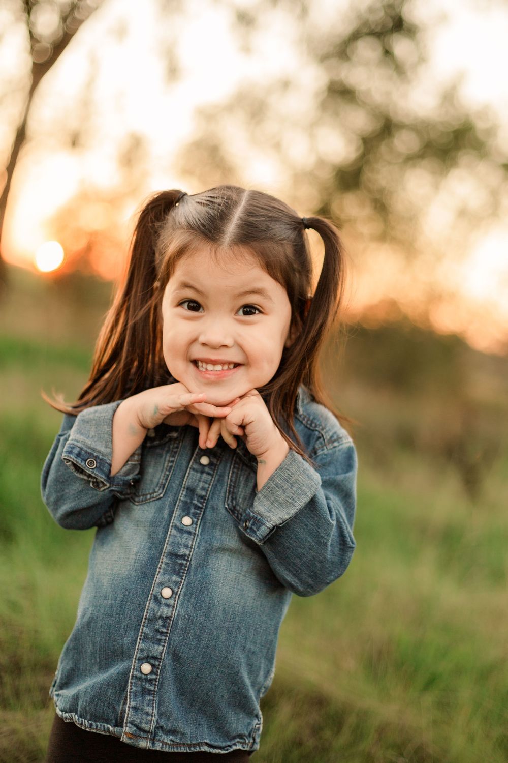A young girl in denim jacket poses with hands under chin against sunset backdrop.