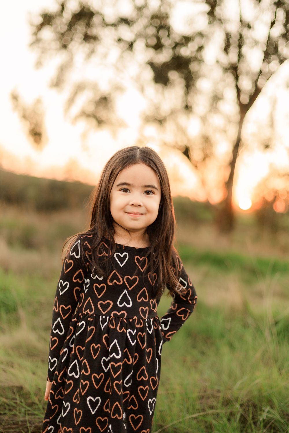 A child wearing a heart-patterned dress stands in a field at golden hour.