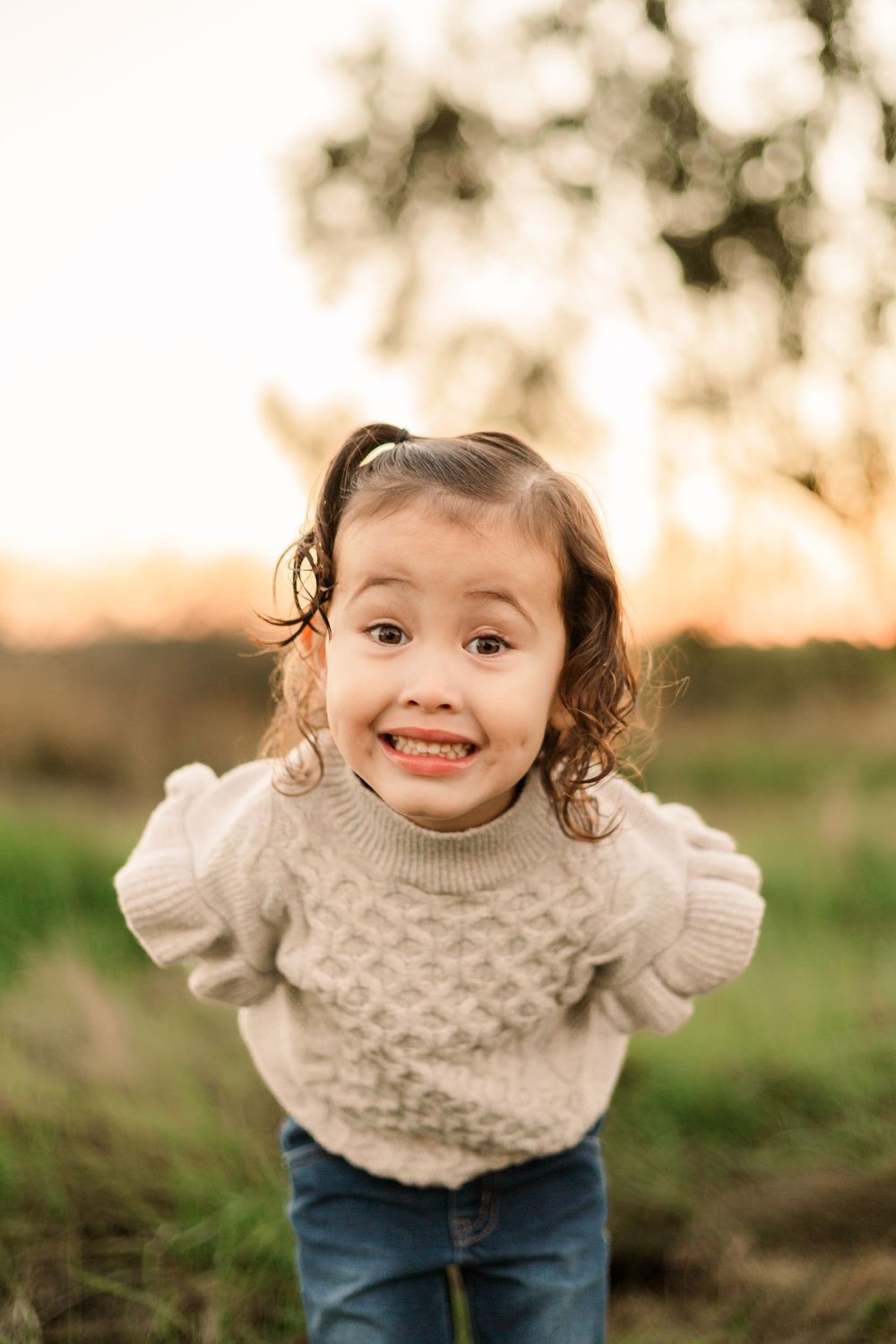 A small child in a cream sweater smiles excitedly at the camera during golden hour.