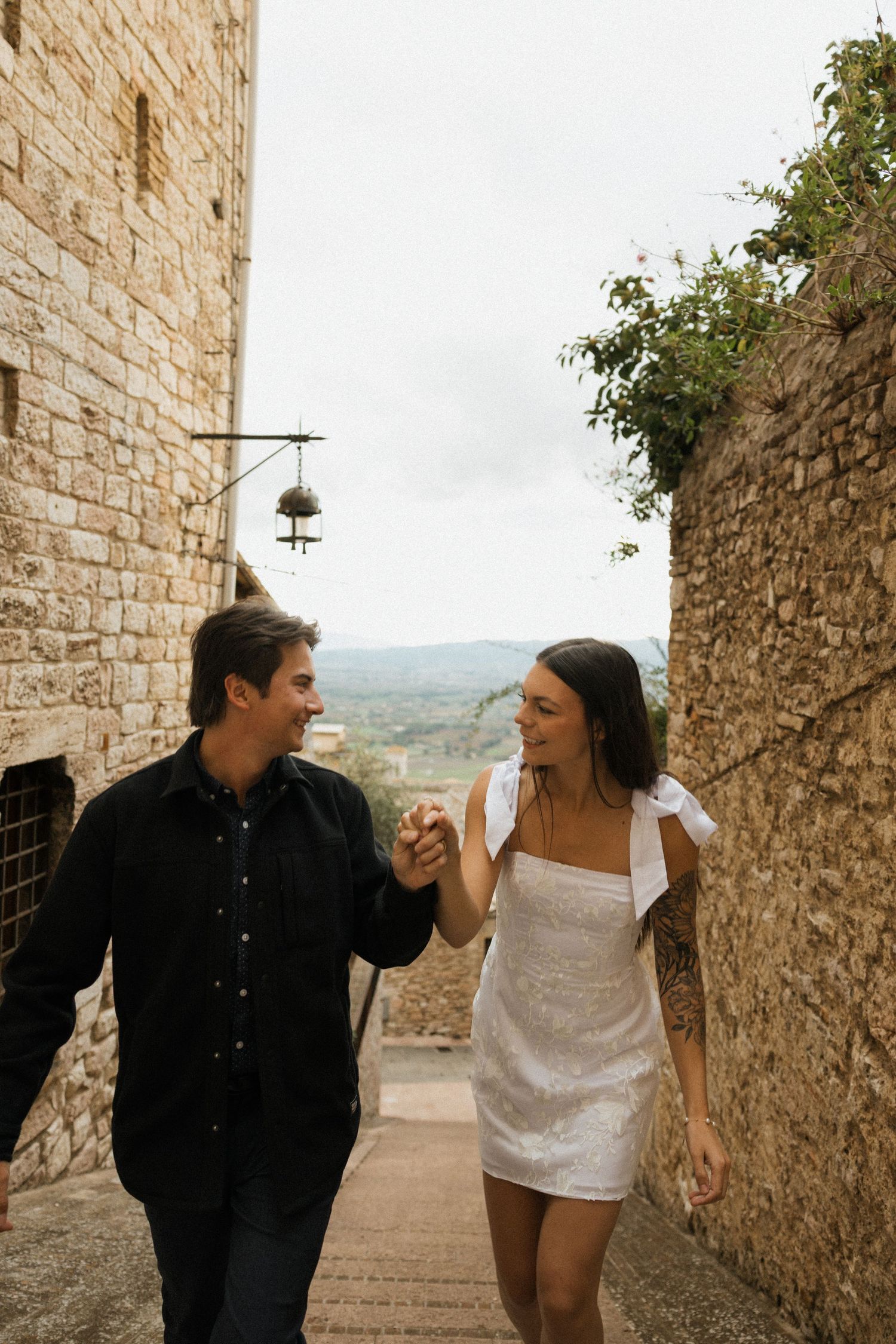 Couple walking and laughing together down rustic stone steps in a Mediterranean village setting.