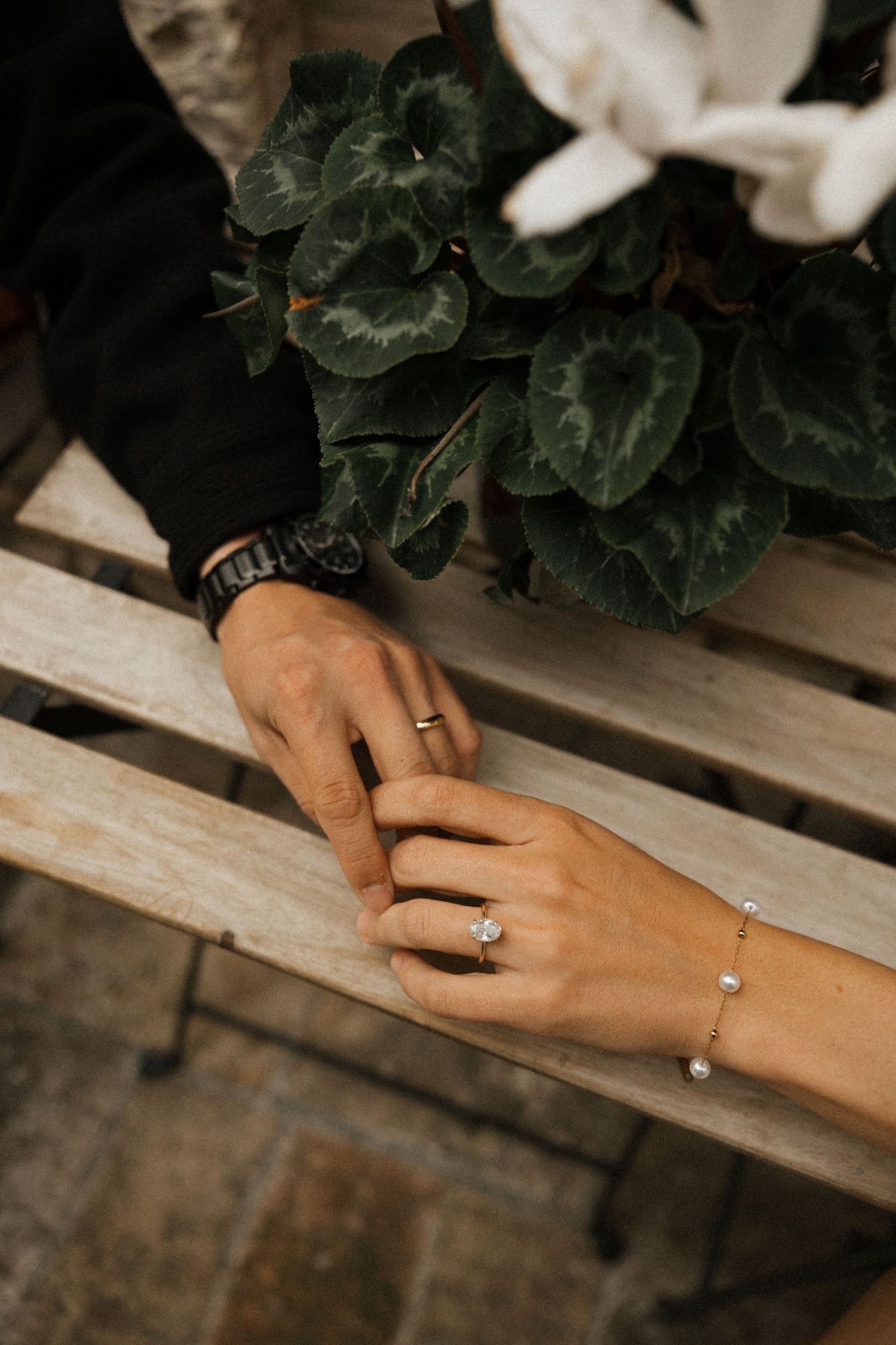 Close up of hands with engagement rings resting on weathered wooden surface next to green foliage.