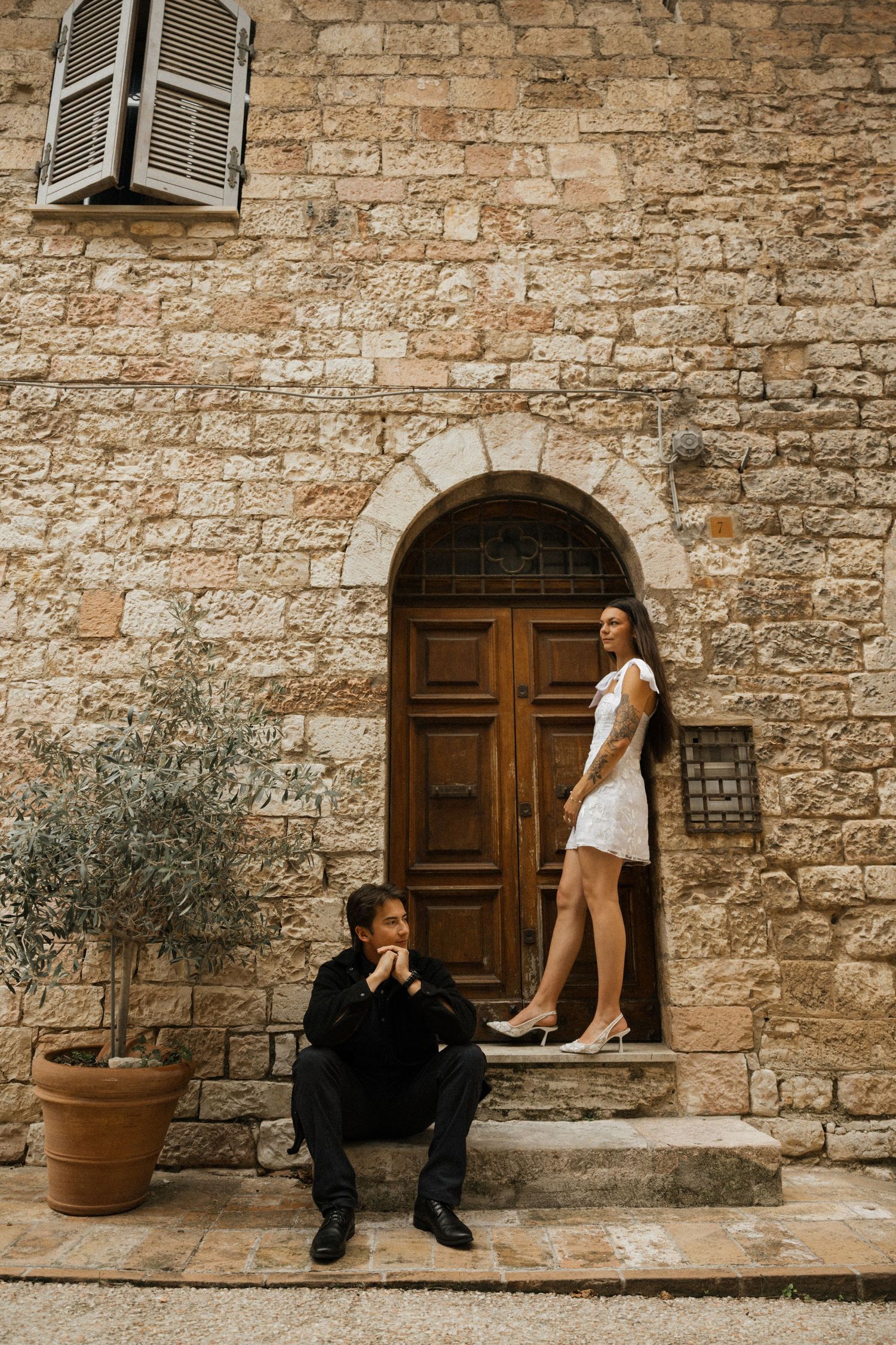 A romantic photoshoot featuring a couple posing against a rustic stone wall with wooden doors and potted plants.