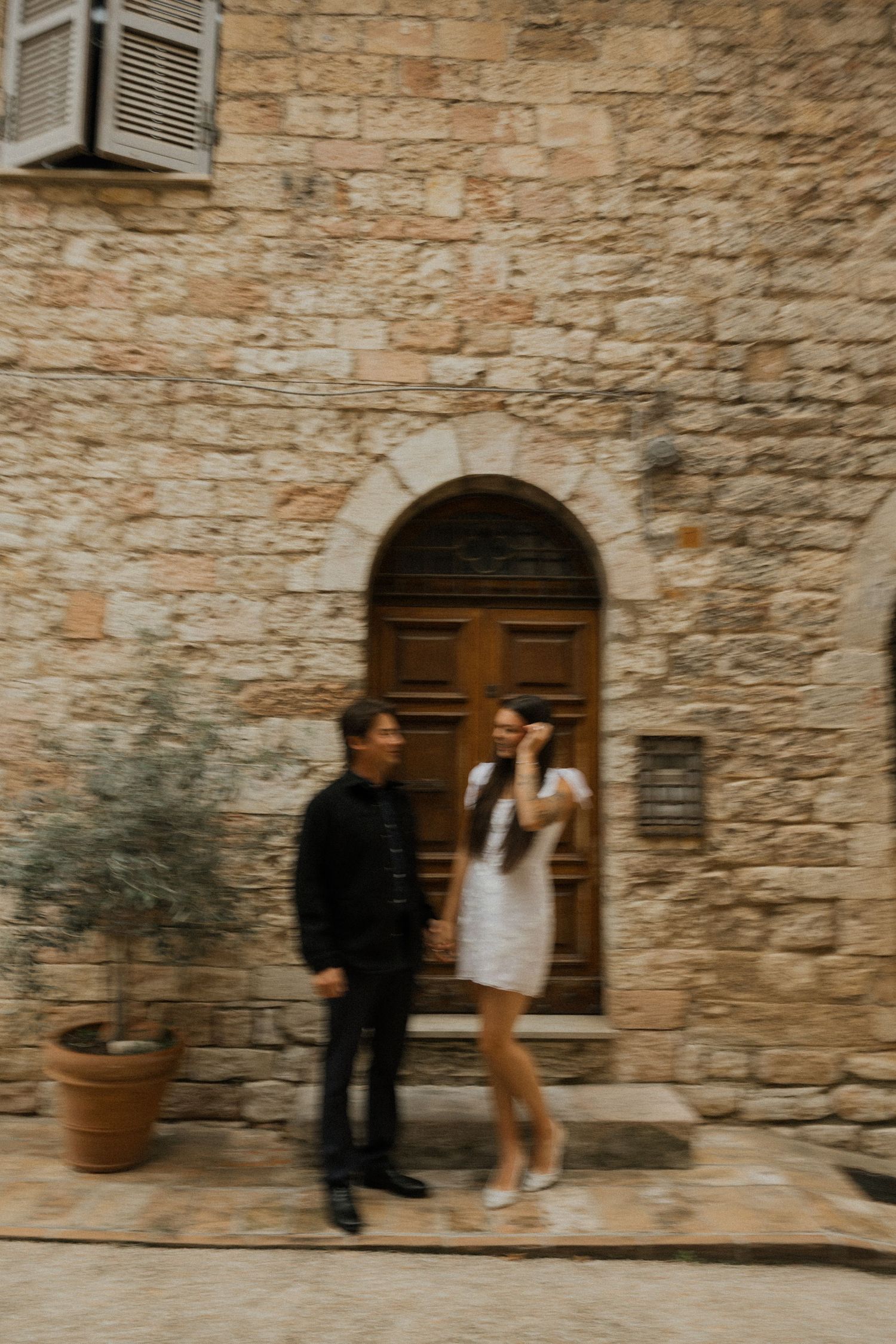 A couple poses against a rustic stone wall with wooden doors in a romantic Italian village setting.