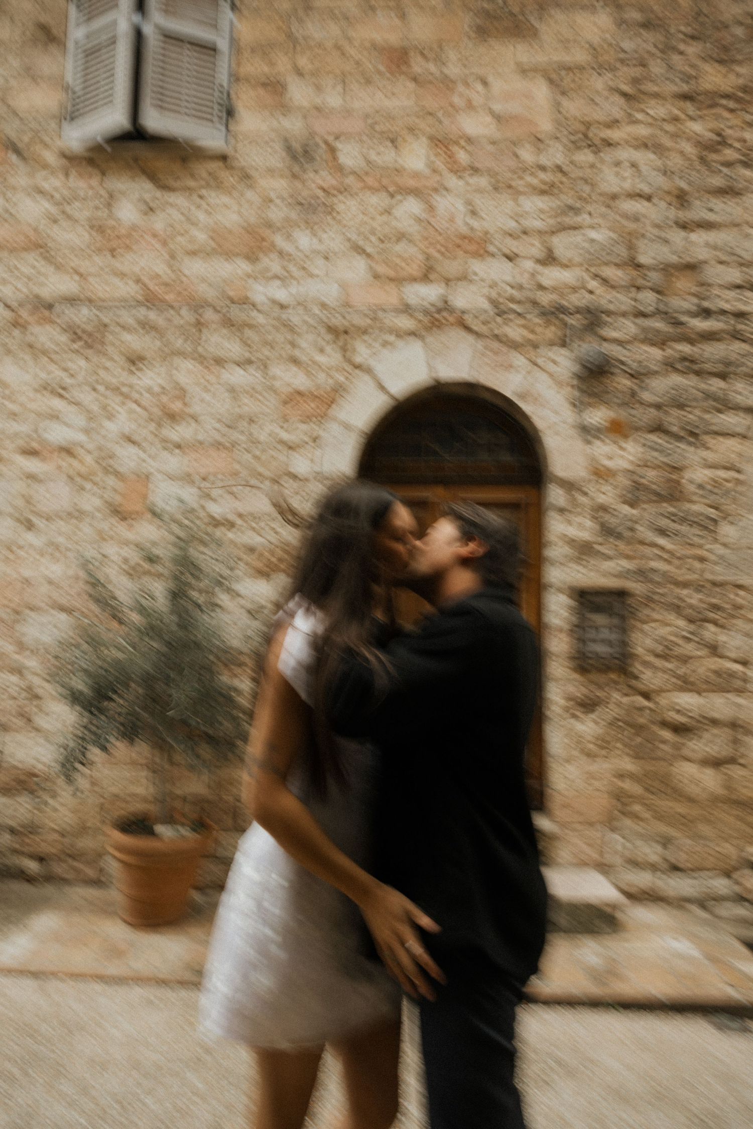 A couple shares a tender embrace against a rustic stone wall in warm, vintage-toned lighting.