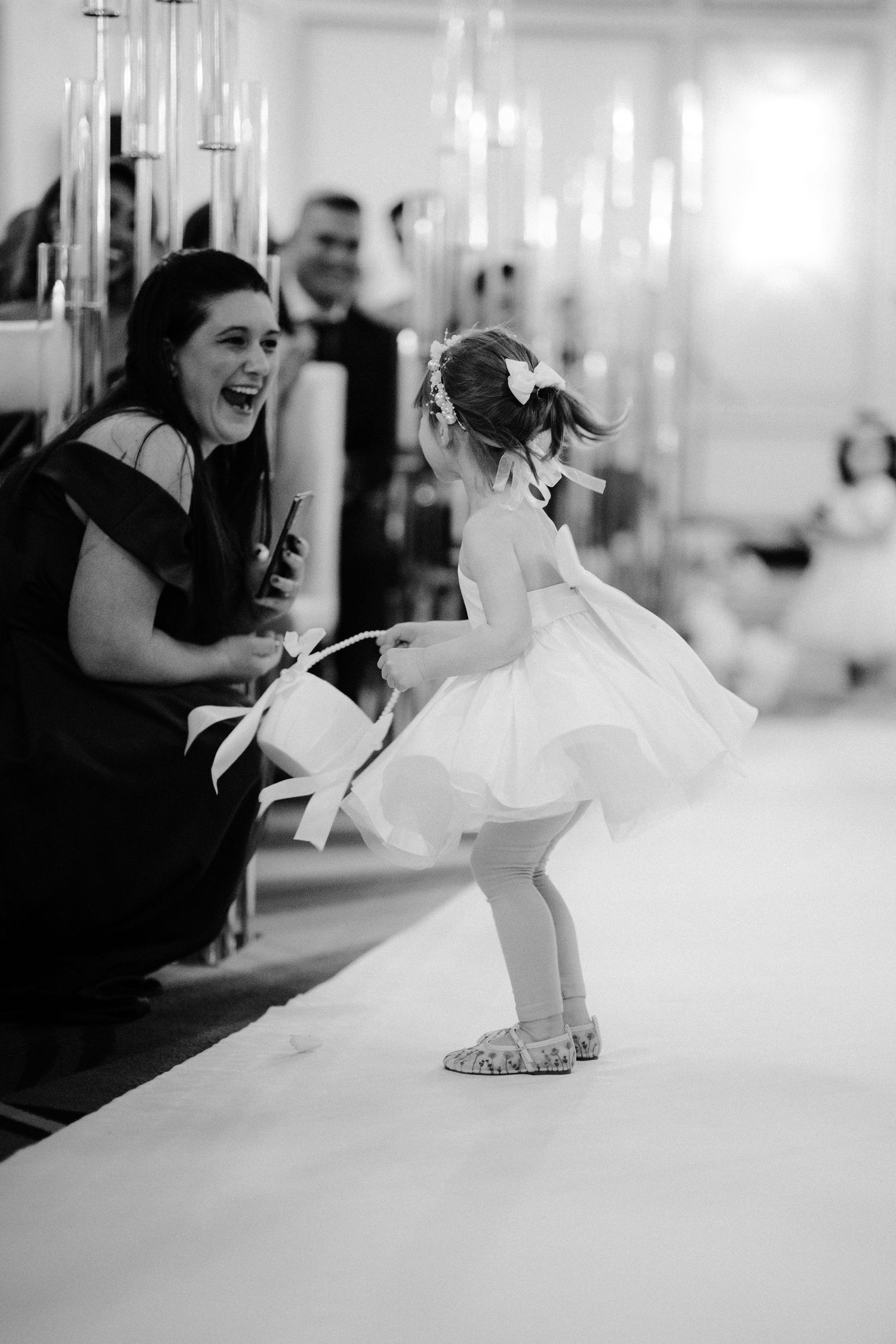 A young flower girl in a white dress walks down a white aisle runner during an elegant wedding ceremony.