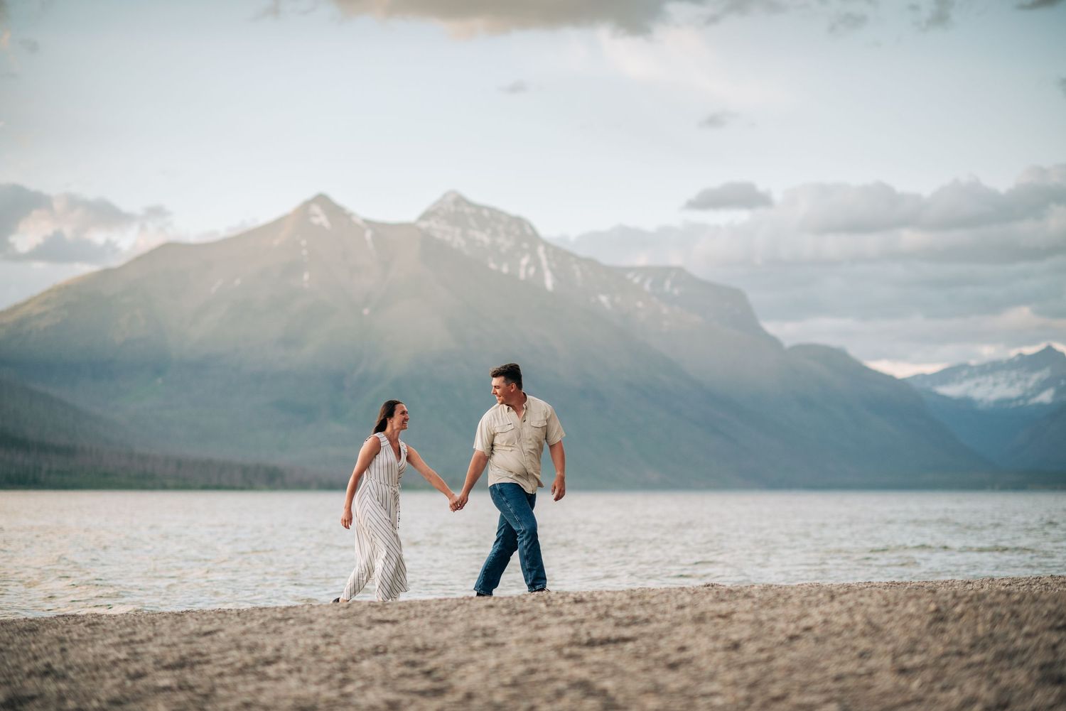 A couple walks together on a scenic beach with majestic mountains and cloudy skies in the background.