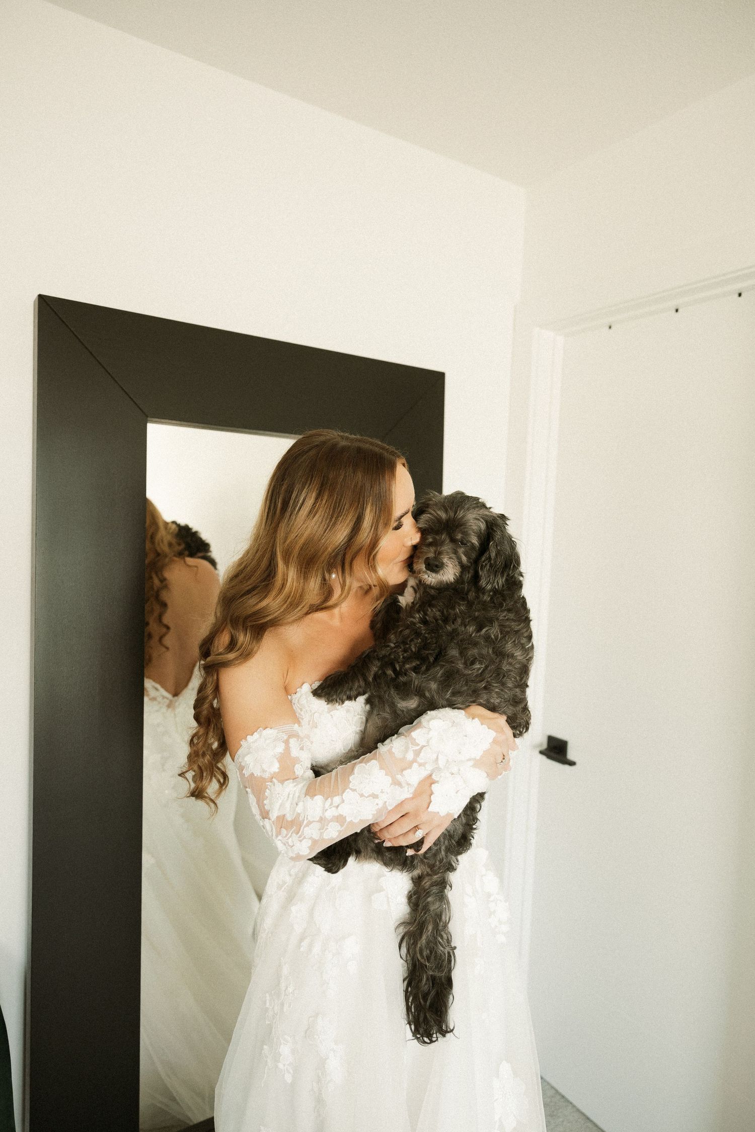 Bride in white lace gown holds black curly-haired dog while standing by mirror.
