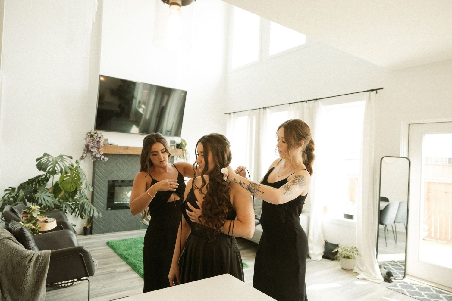 Three people in black dresses getting ready in a bright modern room with a mirror and plants.