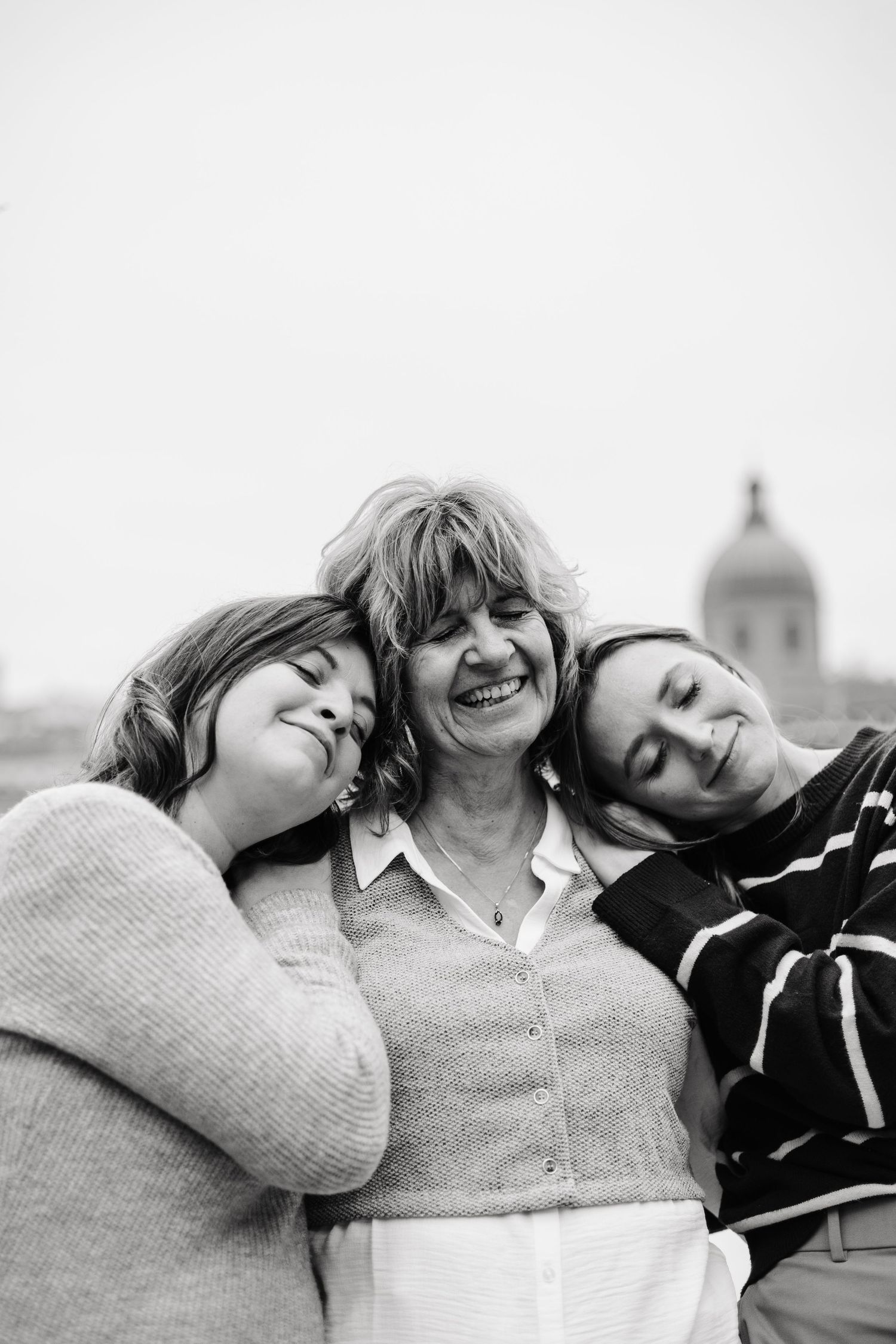 Three women lean together smiling in a black and white photo with Toulouse skyline in background.