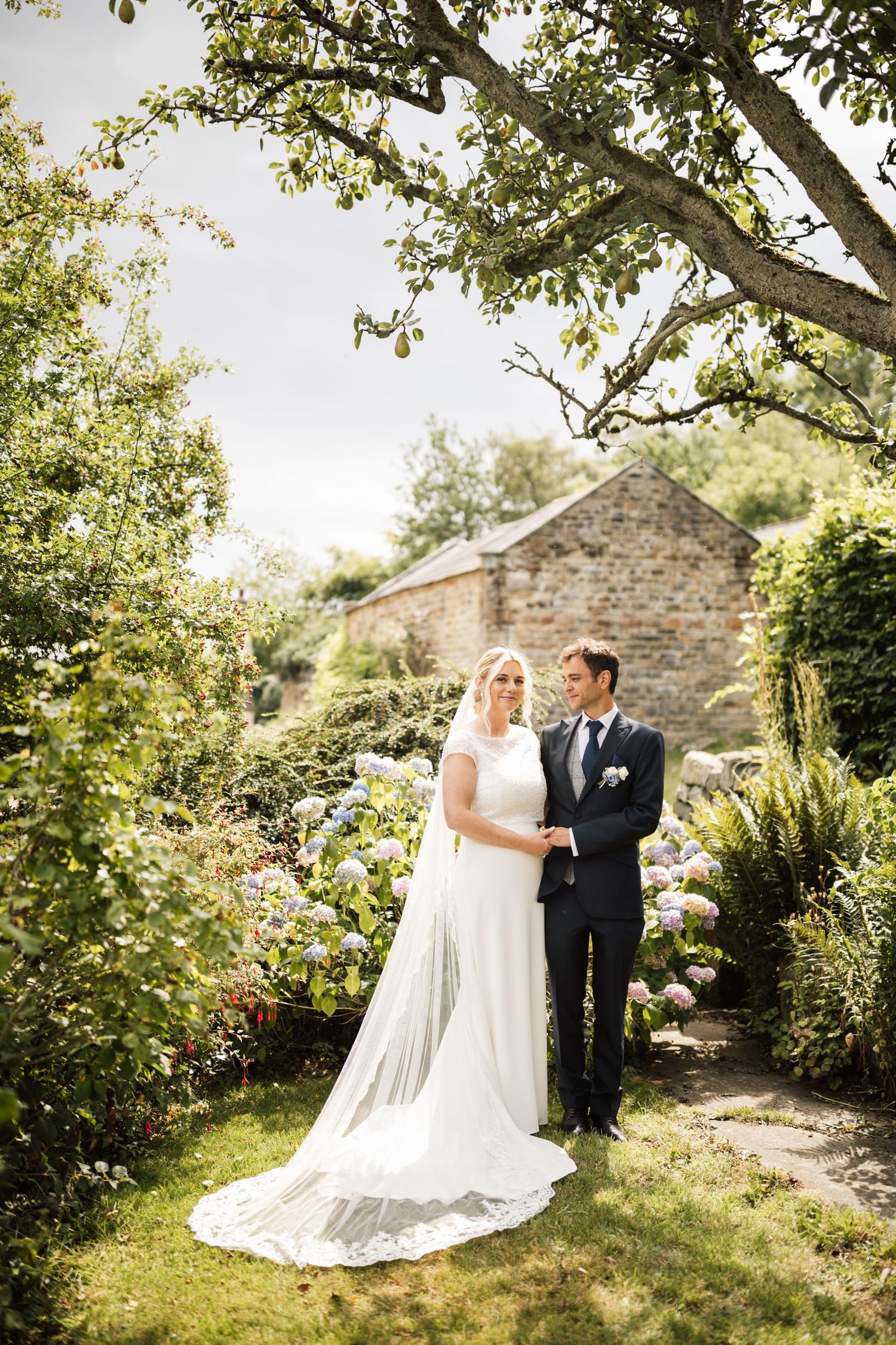 A couple in wedding attire stands together in a sunlit garden setting with stone walls and lush greenery.