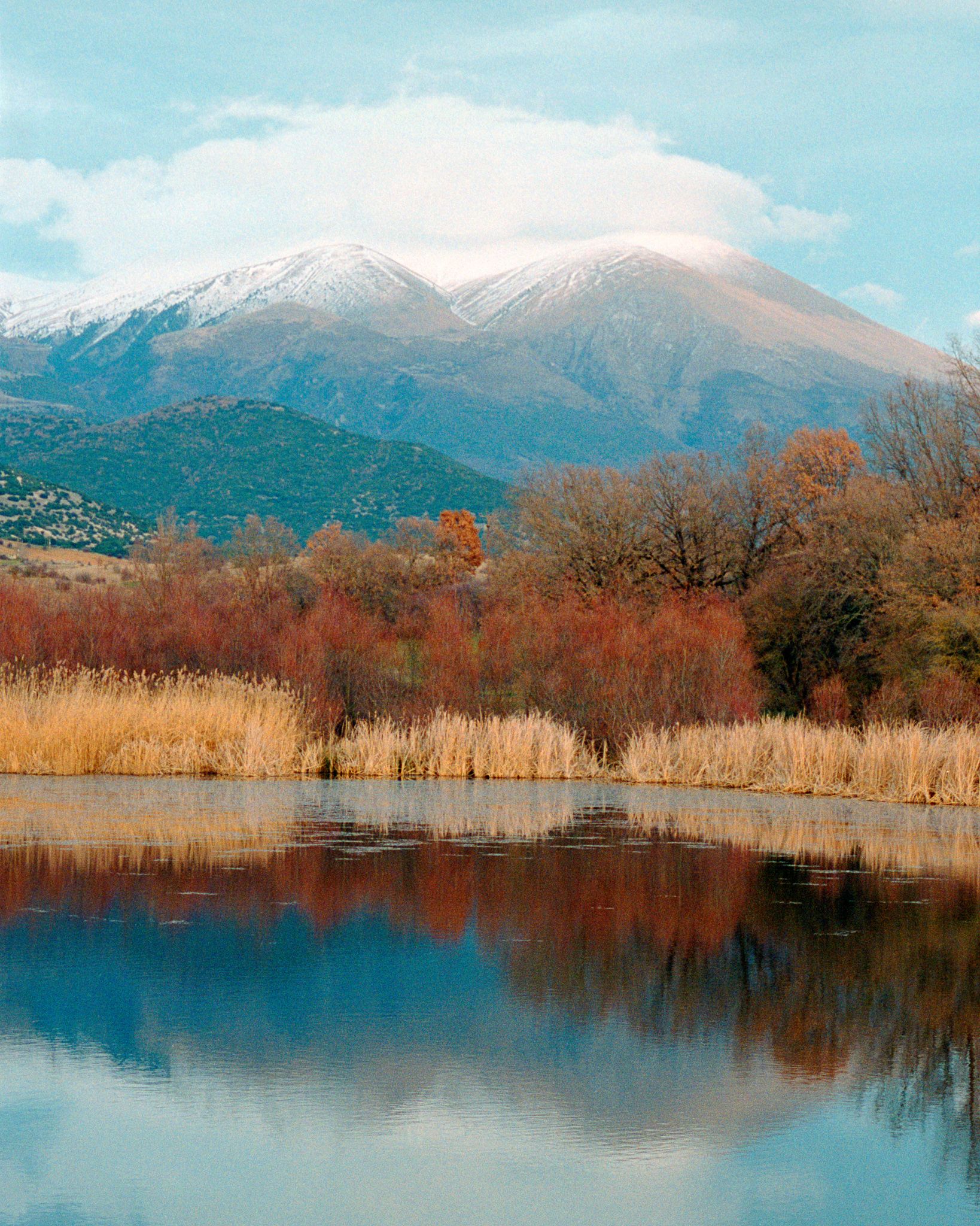 Mountain landscape reflected in calm lake with autumn trees and snow-capped peak.