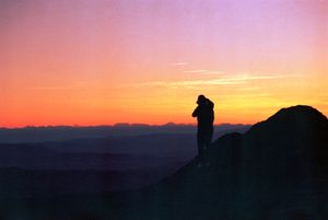 Silhouette of a person standing on mountain peak at sunset overlooking layered landscape.