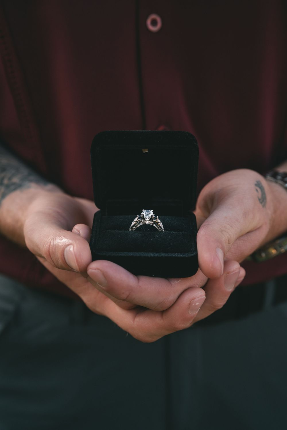 Hands holding a small black ring box with a diamond engagement ring inside.