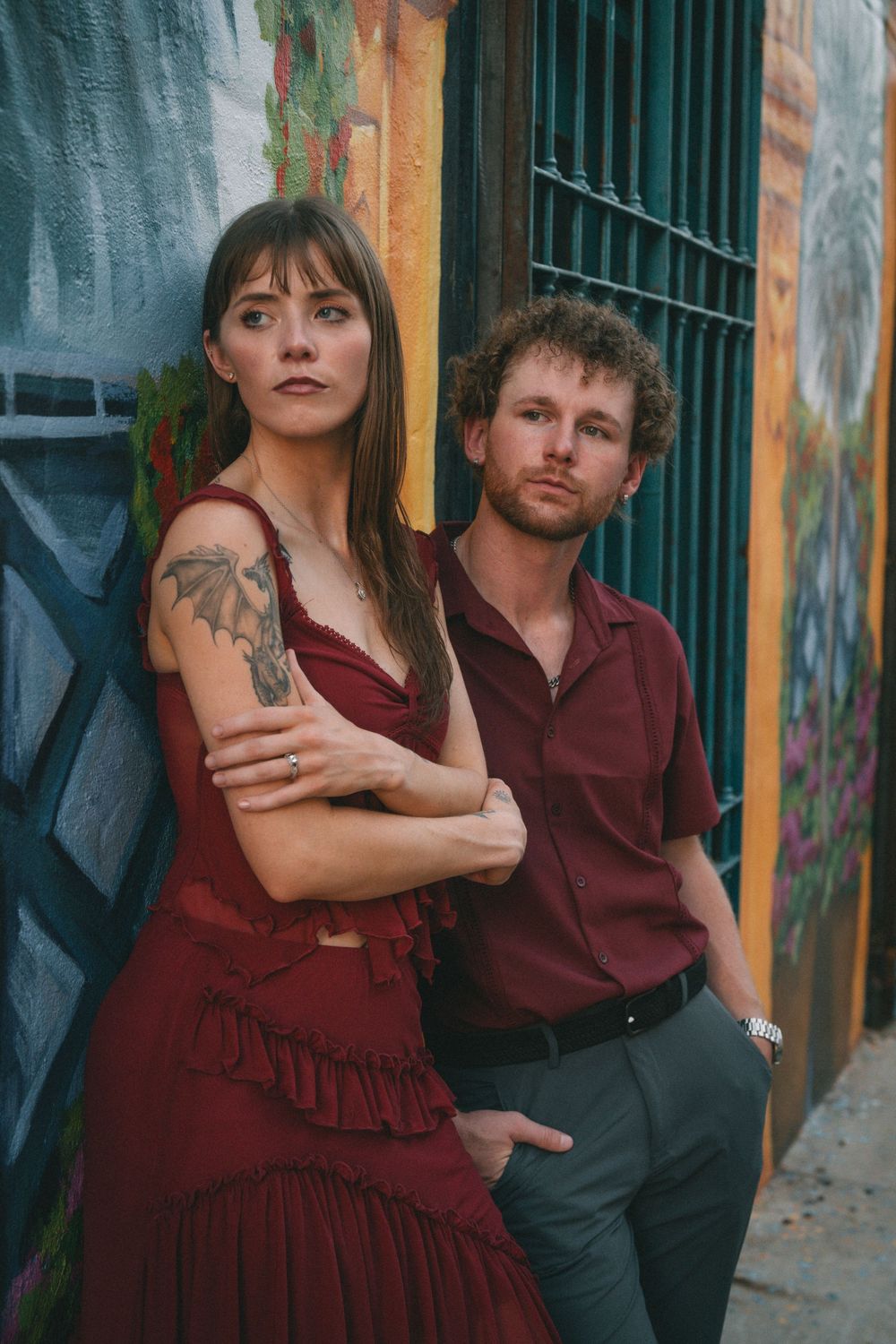 Couple in burgundy and dark clothing pose against a yellow wall with architectural details.