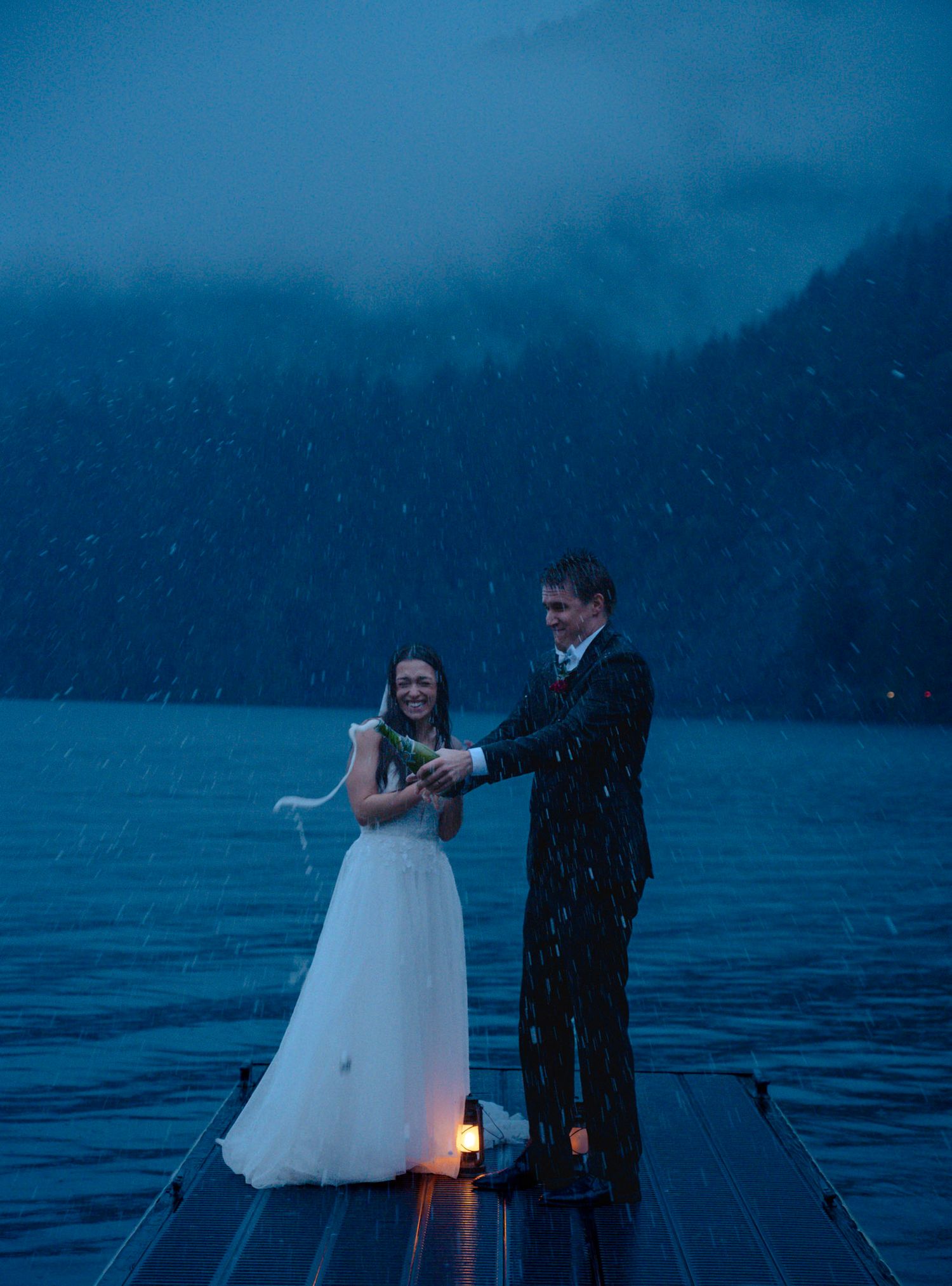 A bride and groom celebrate on a dock in the rain at dusk, with a glowing lantern and misty lake backdrop.