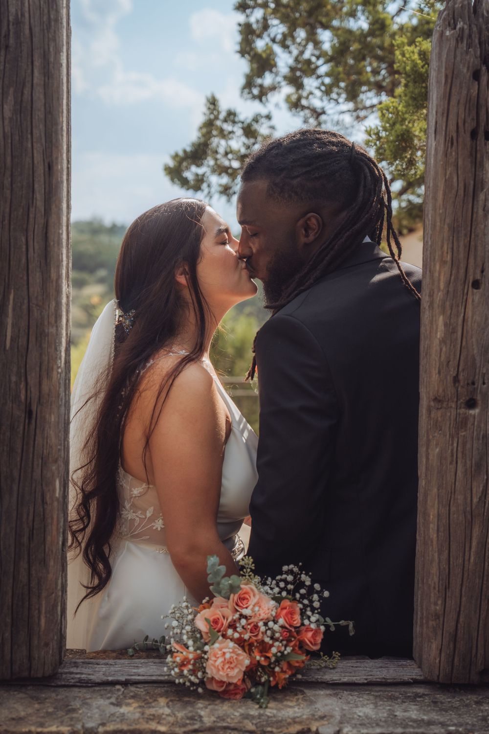 A romantic wedding moment captured in a rustic wooden doorframe with a coral bouquet.