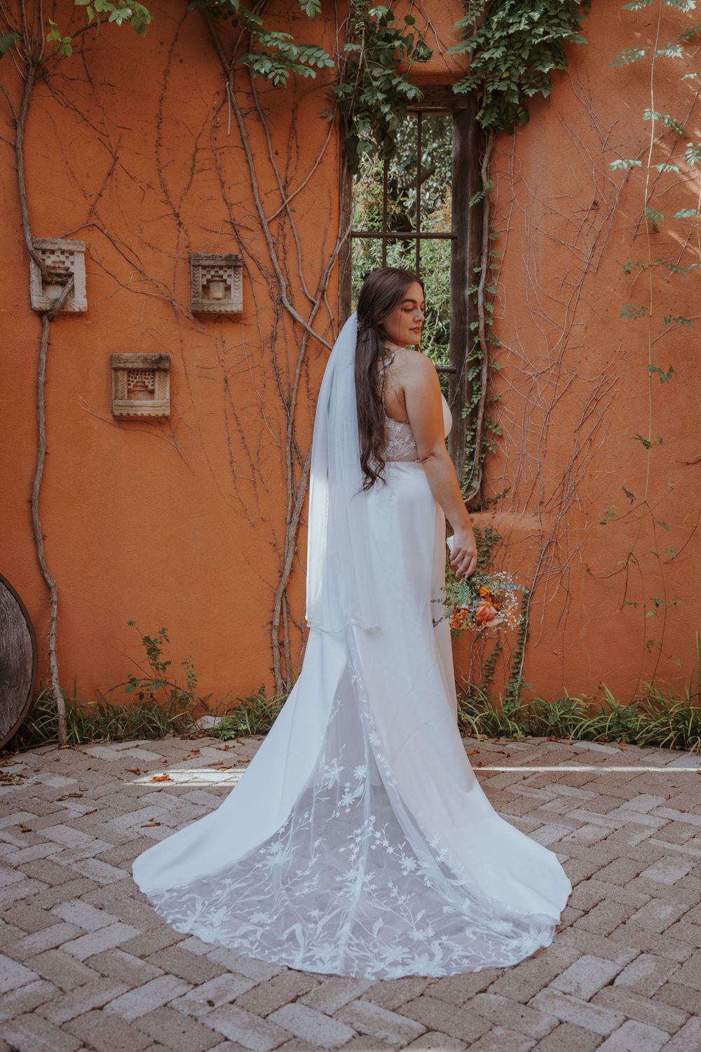 Elegant white wedding dress with lace train flowing across terracotta brick patio near orange wall.