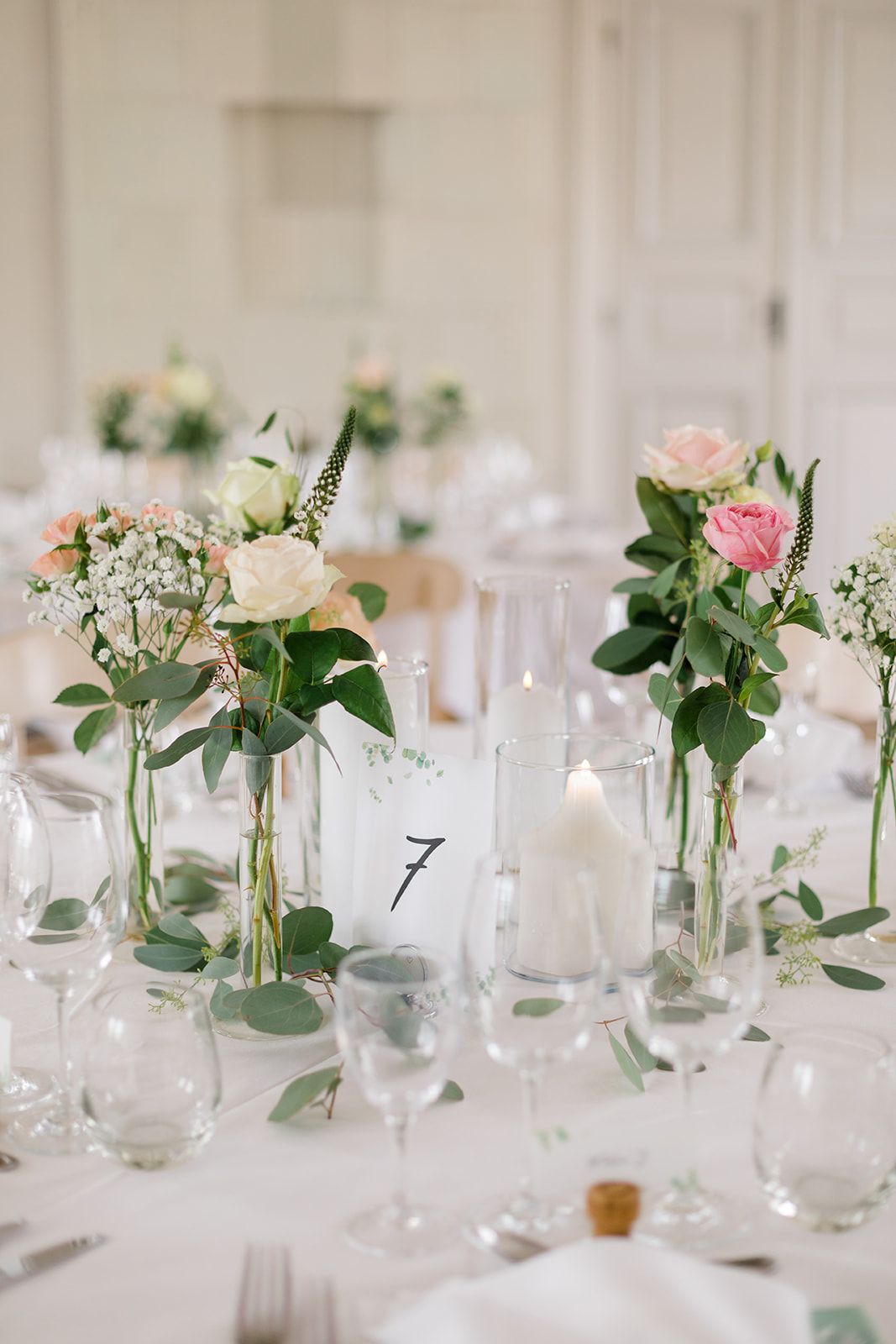 Elegant wedding reception table decor with small glass vases holding pink roses and eucalyptus alongside white candles.