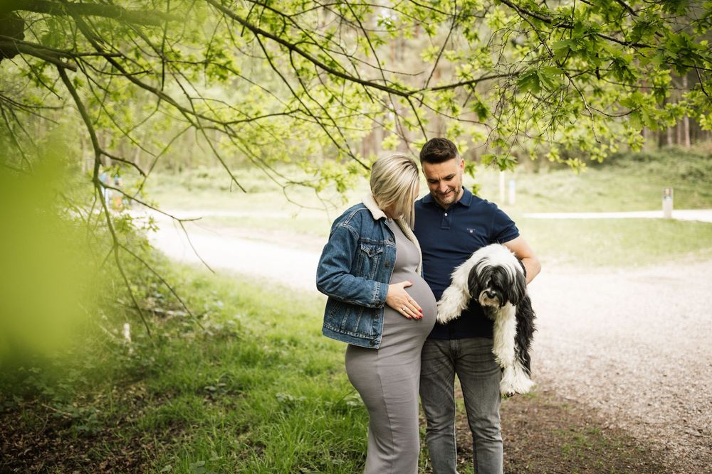 A couple walks with their black dog along a sunlit wooded path during a spring afternoon.