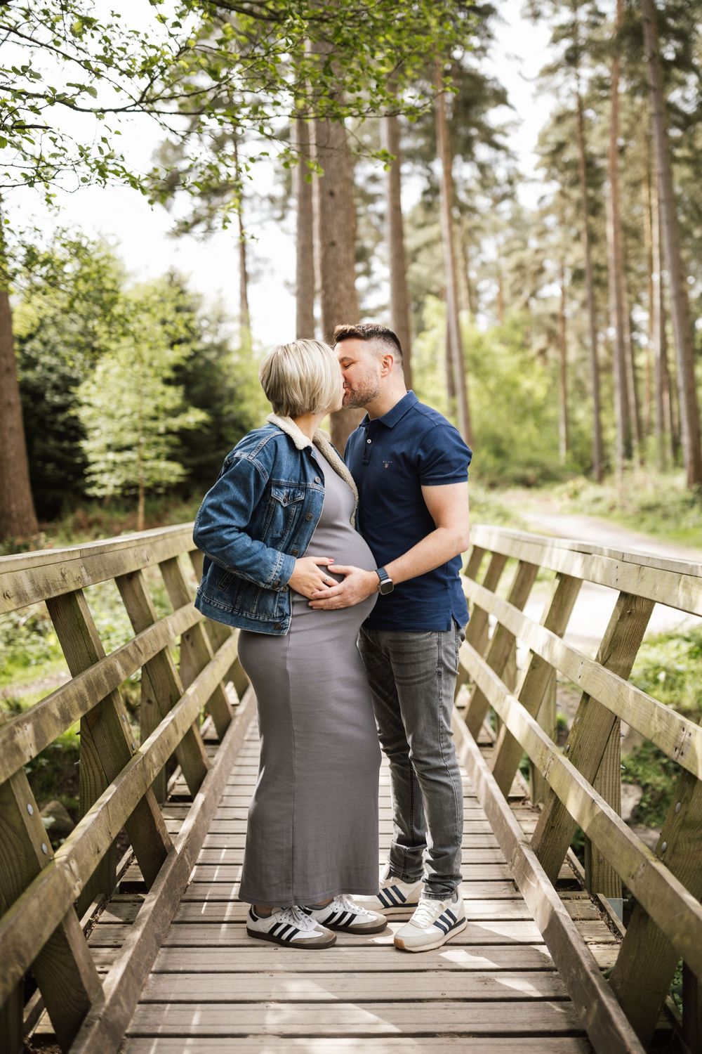 A couple shares an intimate moment on a wooden footbridge surrounded by tall pine trees in a forest setting.
