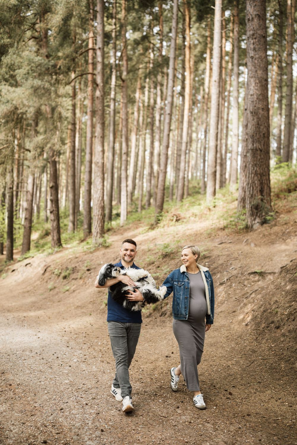 Two people playfully practice martial arts kicks in a sunlit forest setting with tall pine trees in the background.