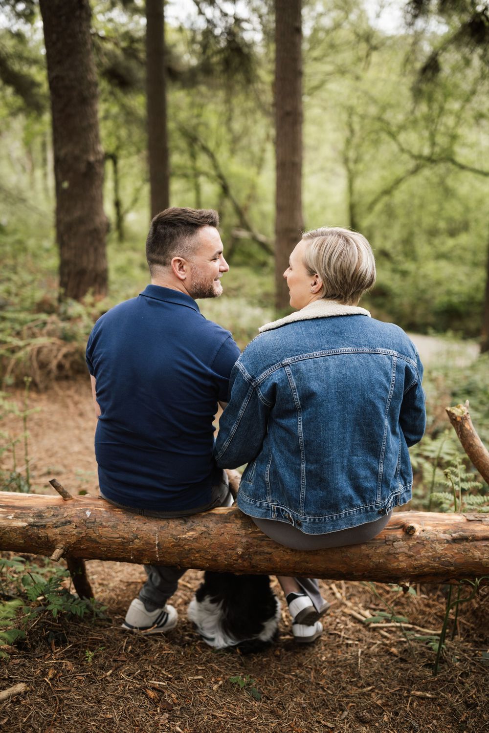 Two people sit together on a fallen log in a peaceful forest setting wearing denim and navy clothing.