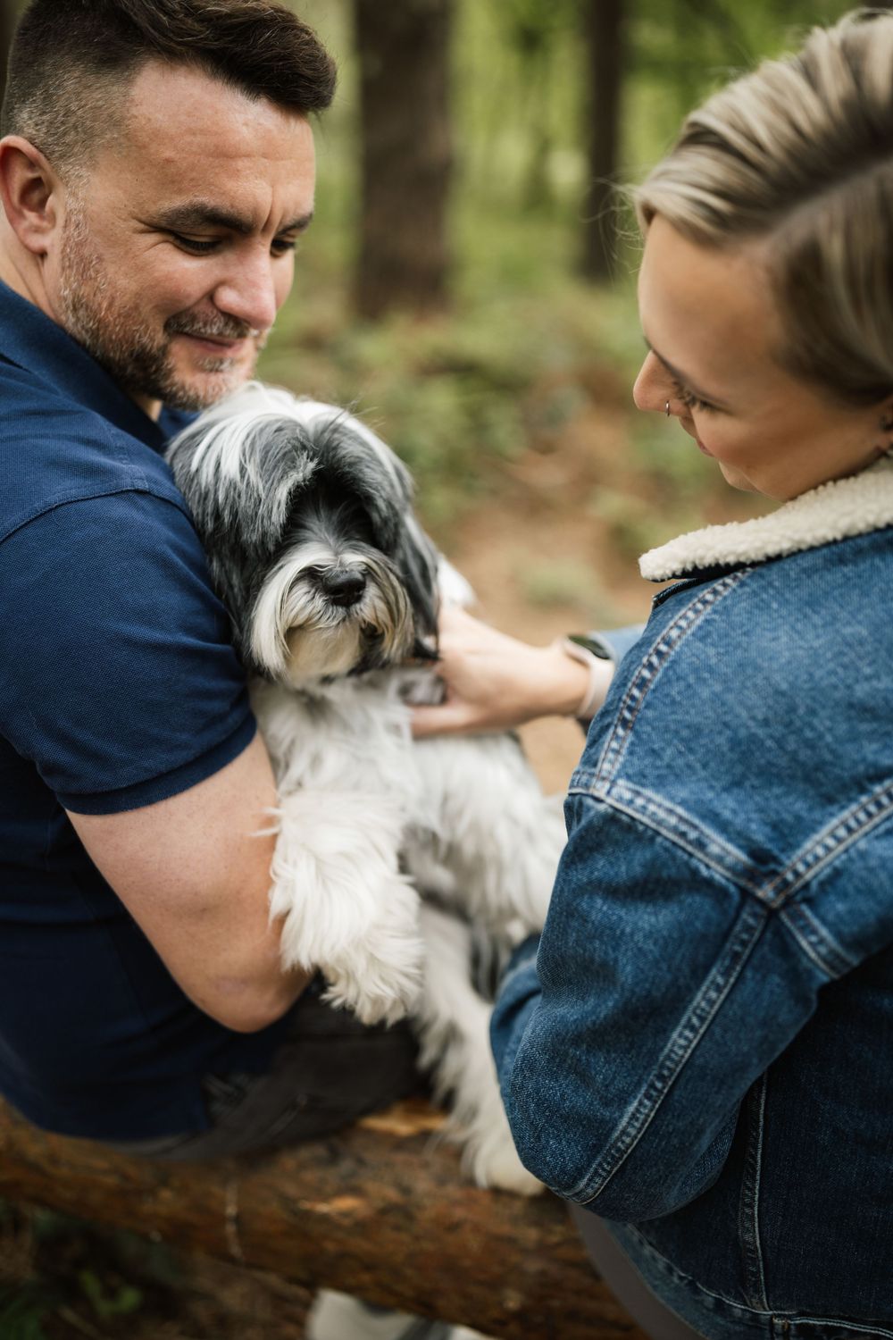 A couple sits on a fallen log in the forest with their small white dog in a sweet candid moment.