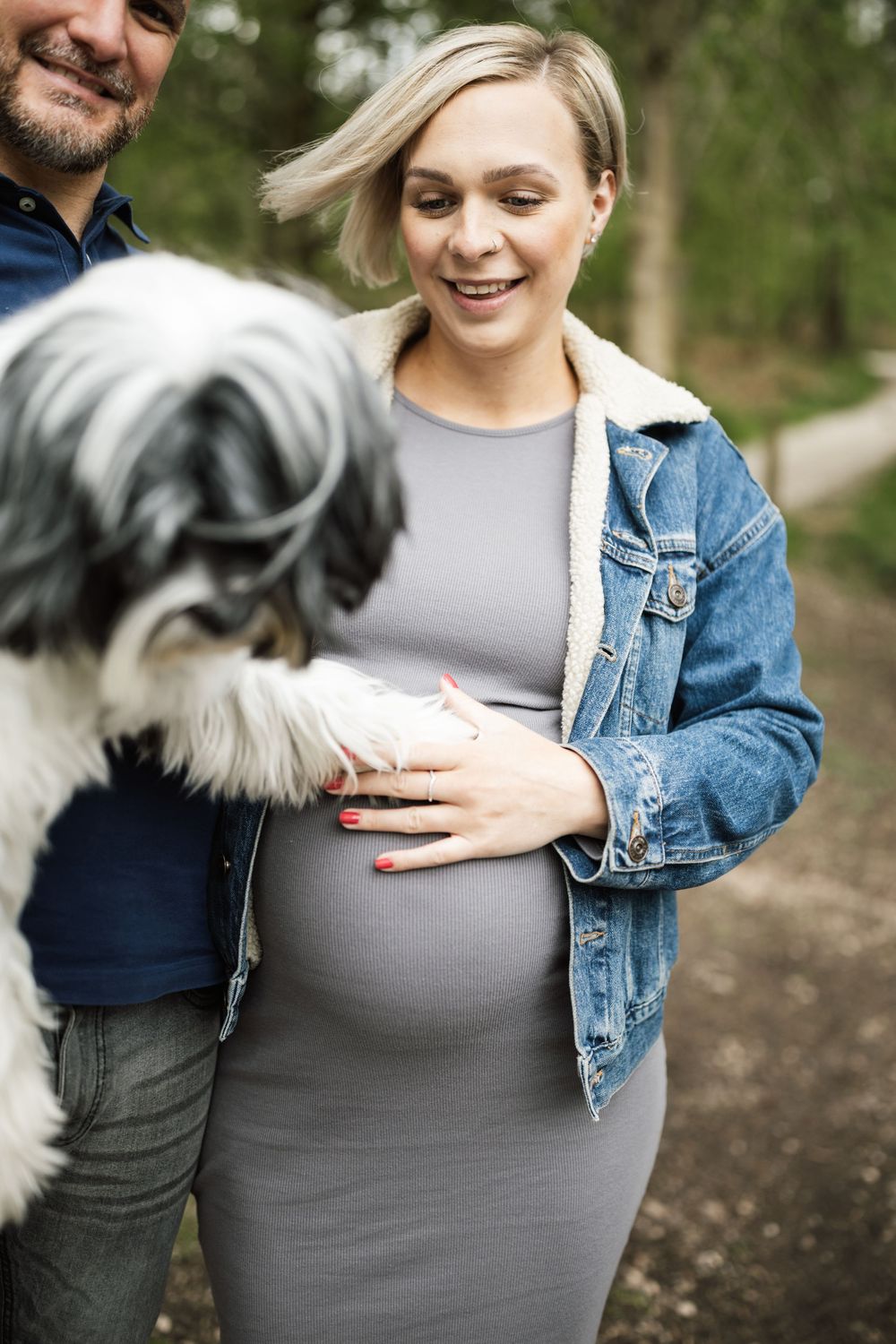 A pregnant woman in a denim jacket poses with her dog while her partner touches her baby bump in the forest.